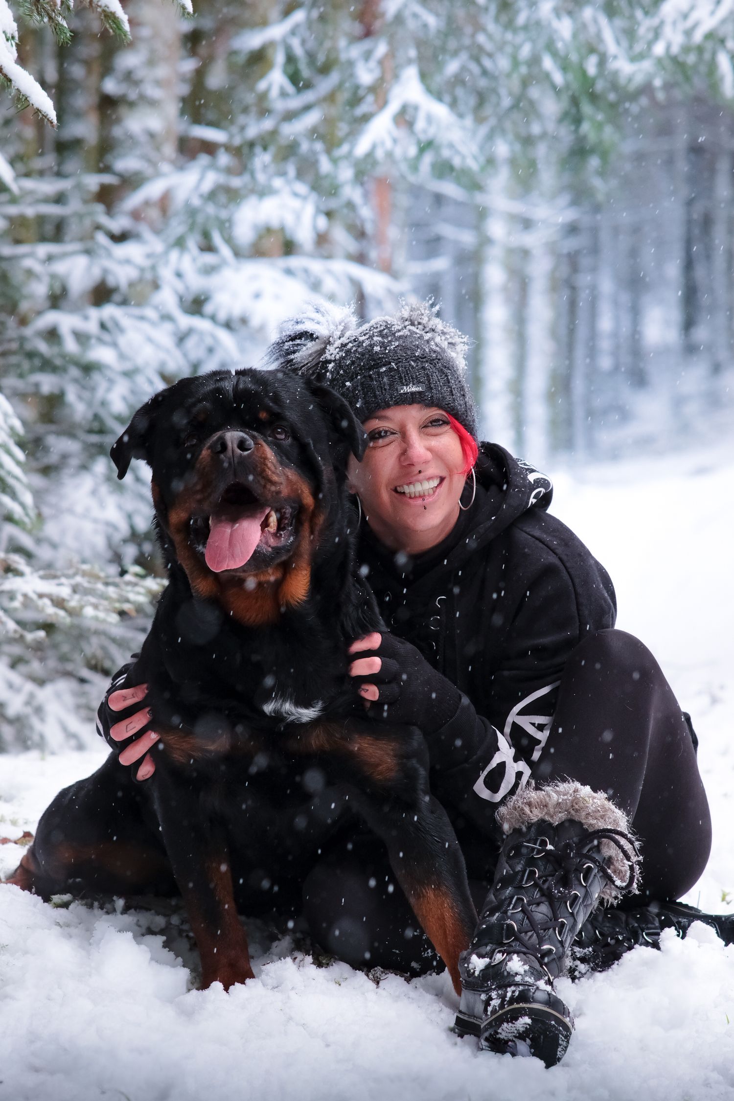 Smiling woman hugging her Rottweiler dog in a snowy winter forest during snowfall.