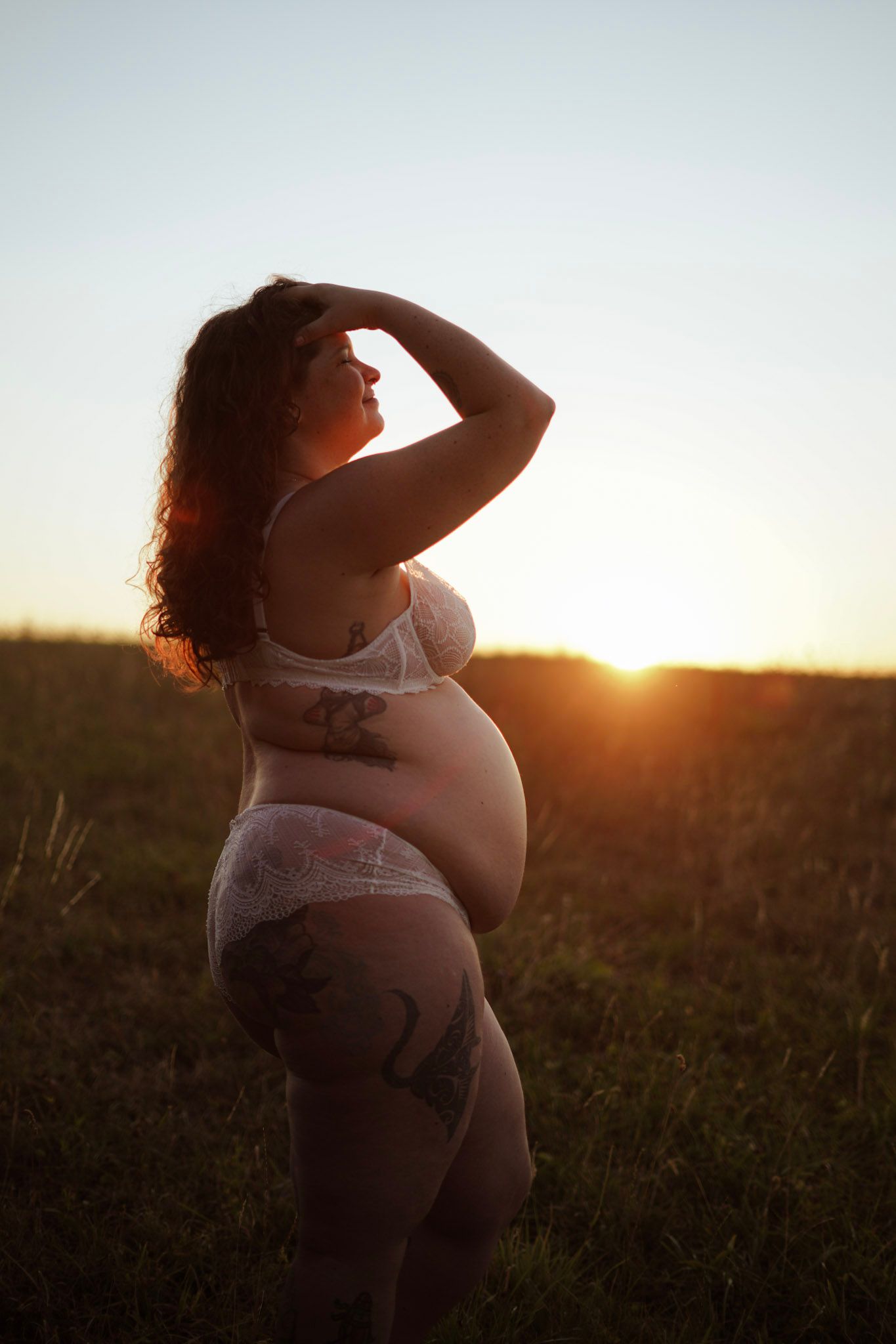 Pregnant woman with tattoos in white lace lingerie posing in a field during golden sunset.