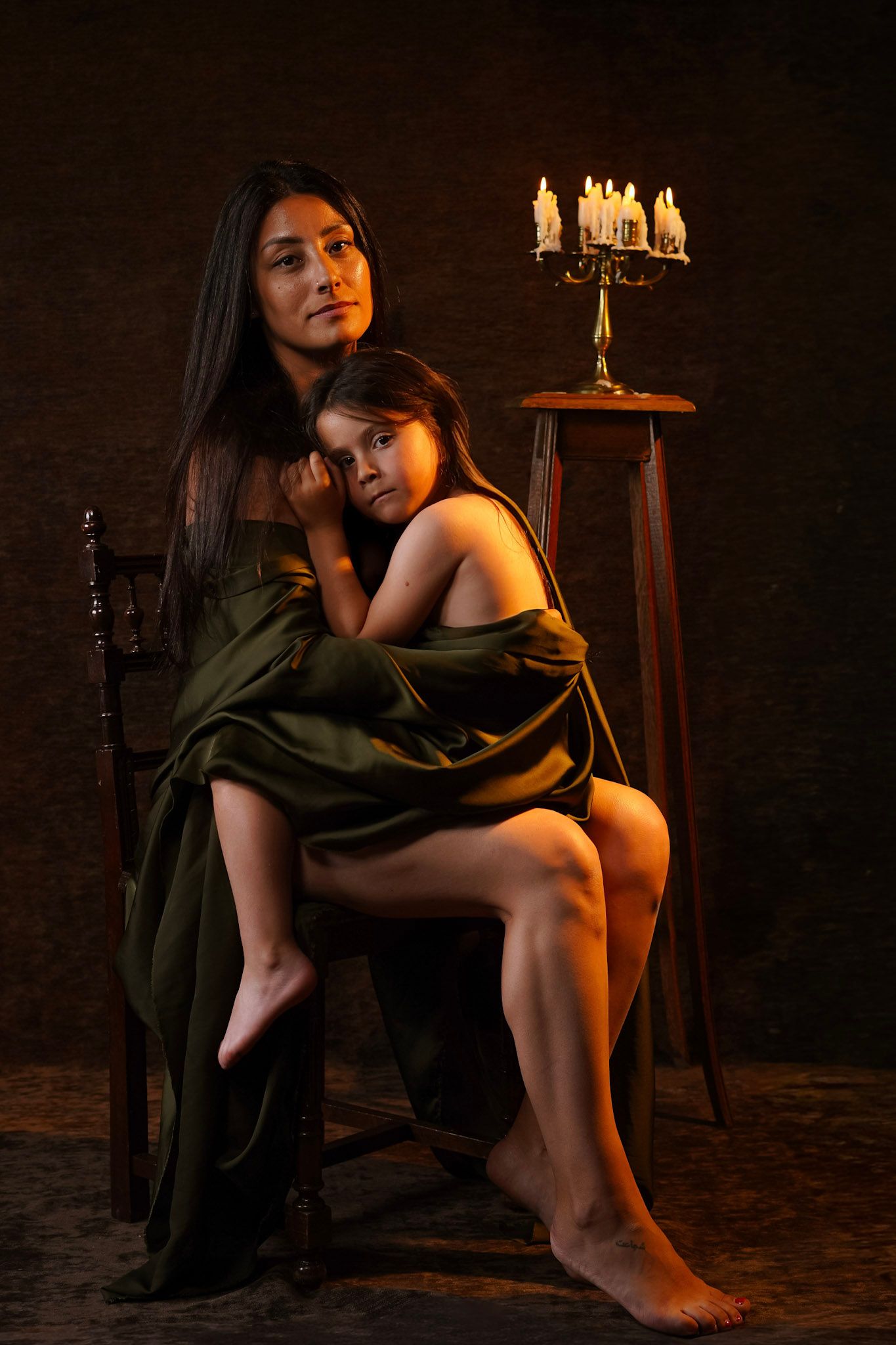 Mother and daughter wrapped in green fabric posing by candlelit candelabra in studio.
