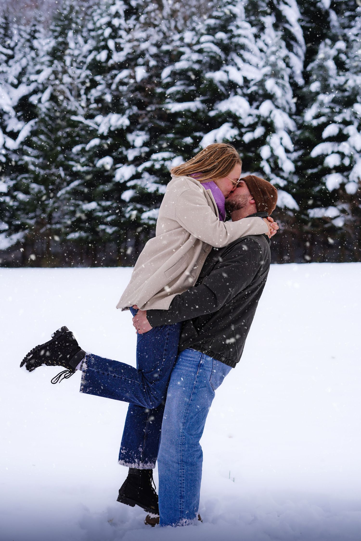 Couple kissing in a snowy winter scene with snow-covered pine trees behind them.