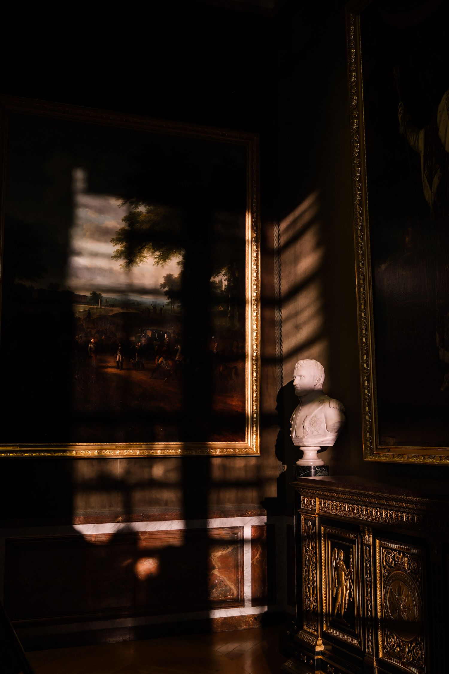 Dramatic chiaroscuro interior with golden-framed painting, marble bust on ornate cabinet, and sunlight casting geometric shadows.