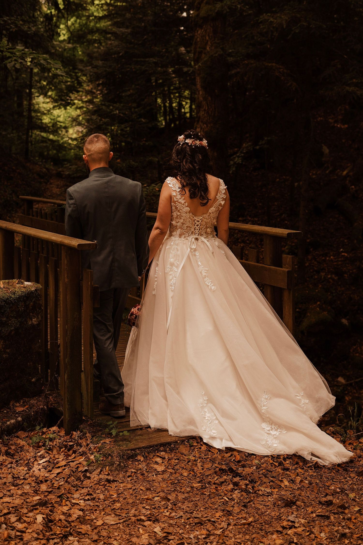 Bride and groom walking hand in hand on a wooden bridge surrounded by autumn forest foliage.