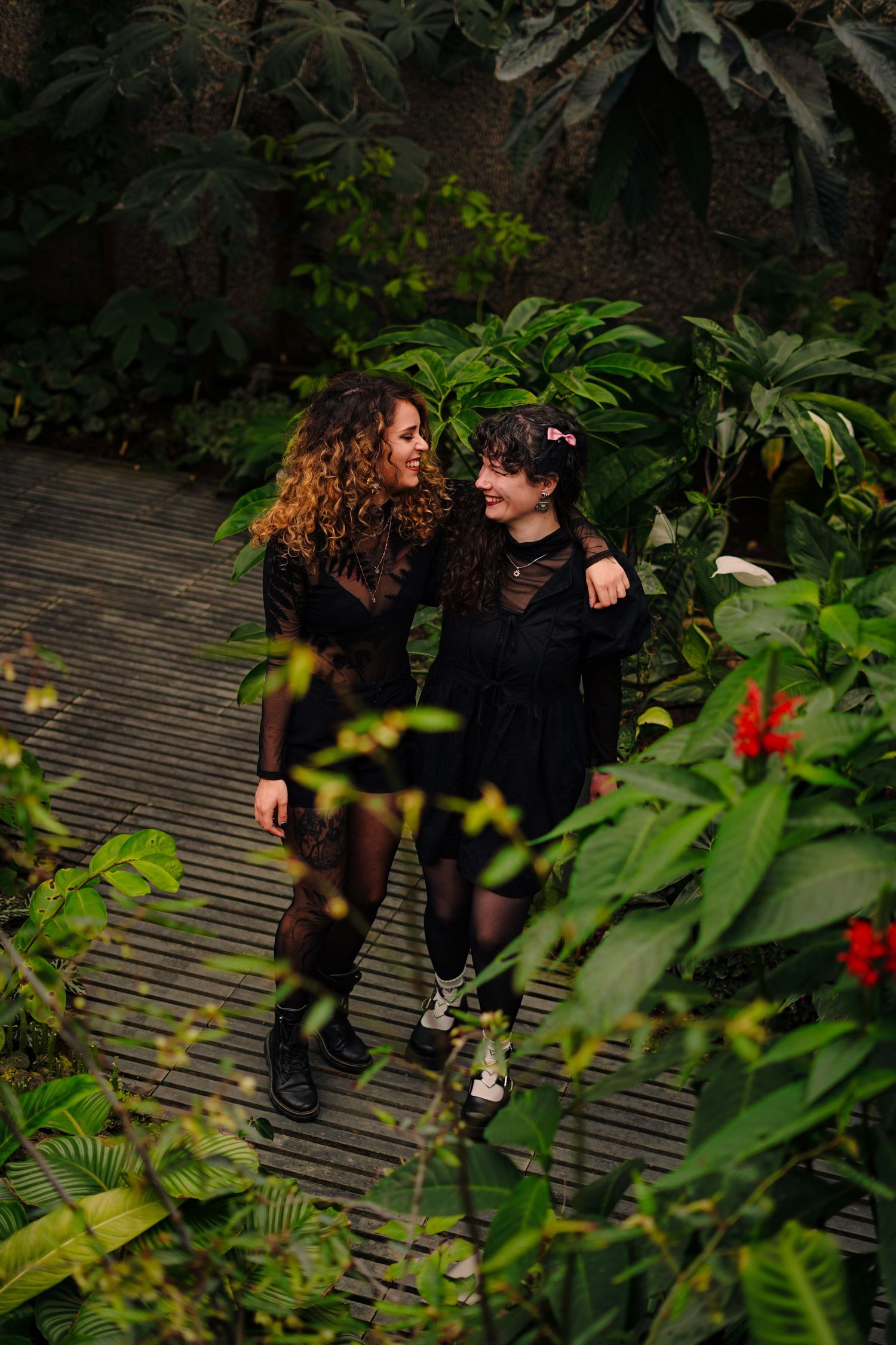 Two women in black dresses laughing together in a lush tropical garden setting.