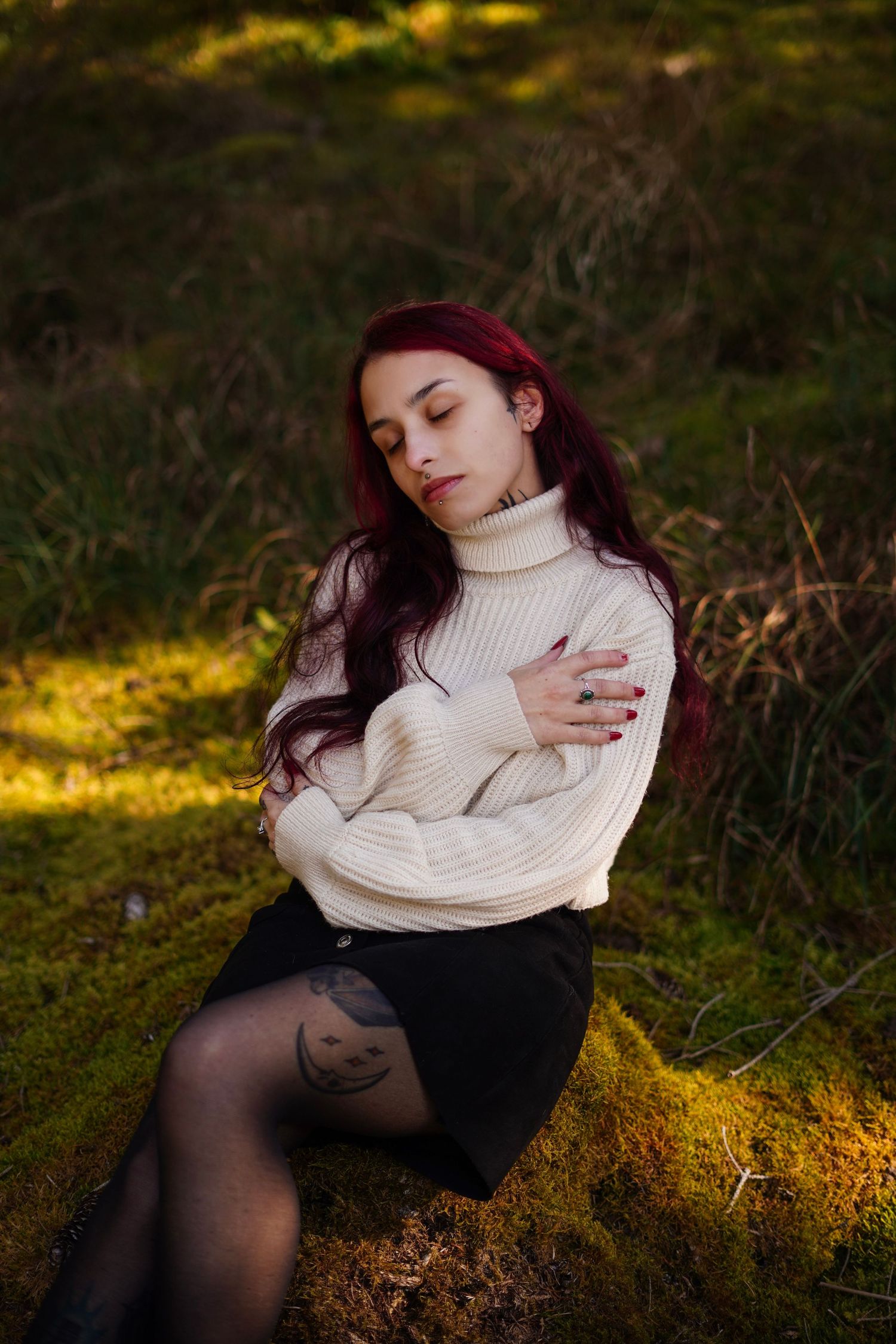 A red-haired woman in a cream sweater and black skirt sits on mossy ground in dappled forest light.