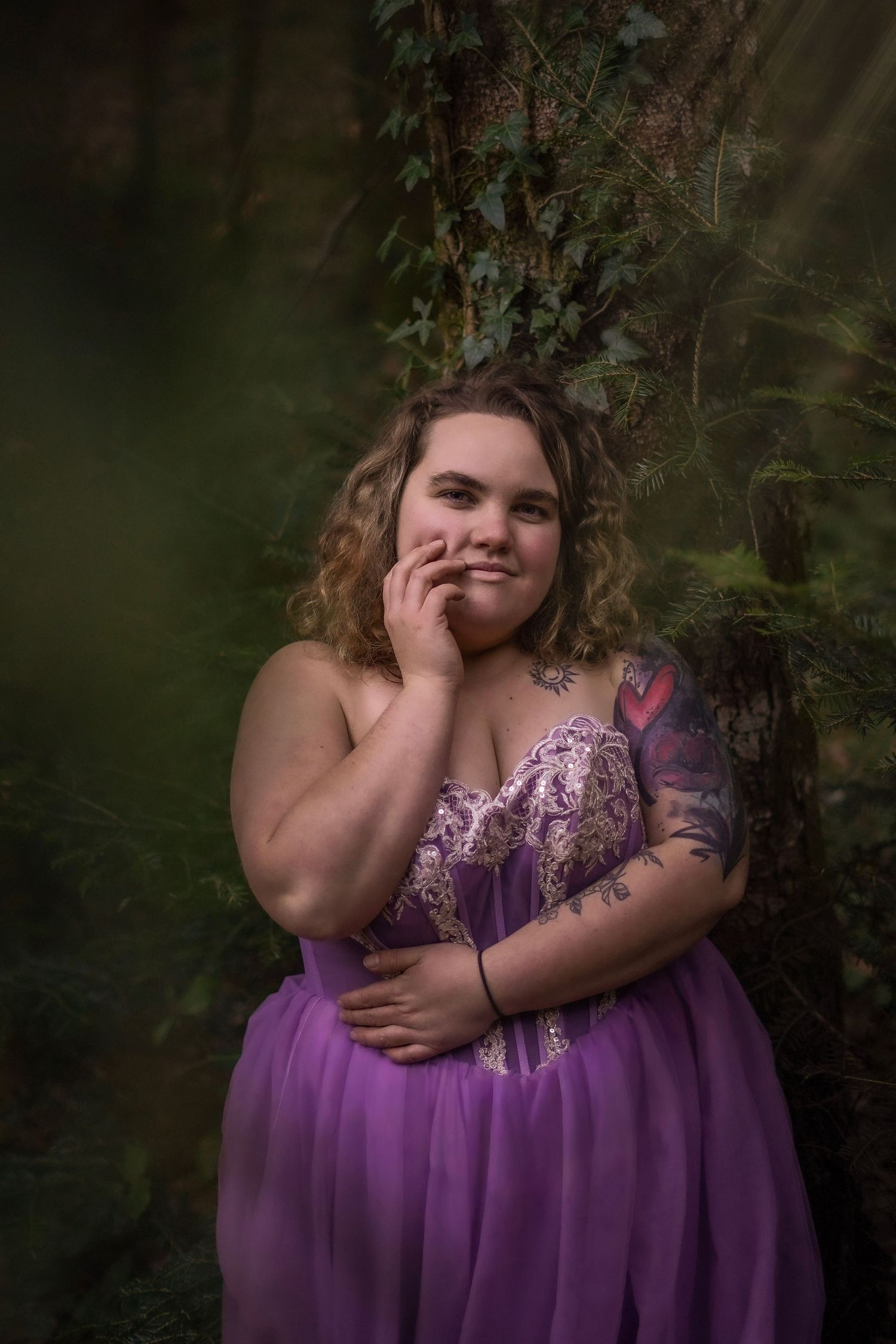 A plus-size woman in a purple gown poses gracefully against ivy-covered trees in an enchanted forest.