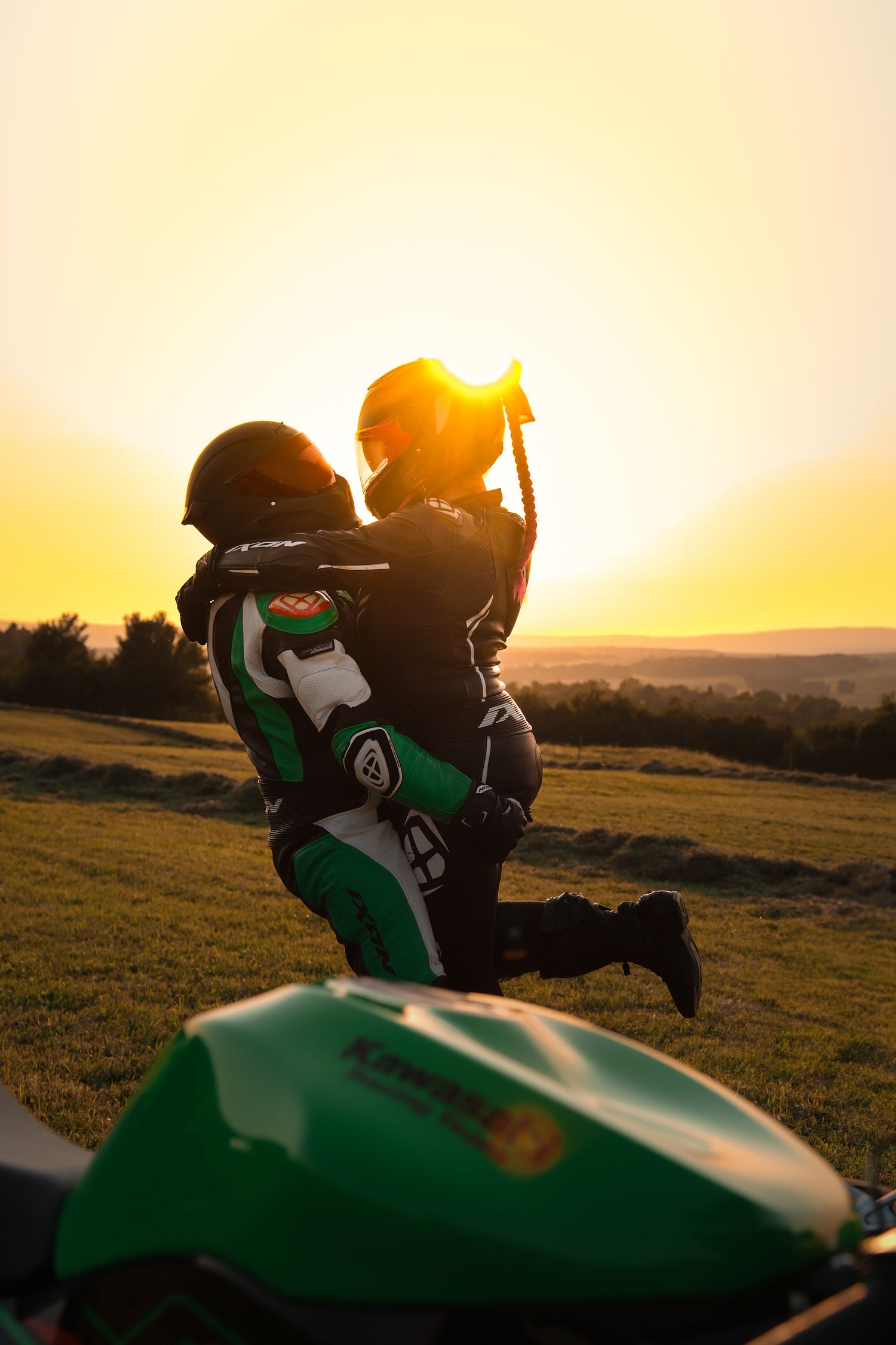 Two motorcyclists in racing gear embracing at golden sunset beside green Kawasaki bike.