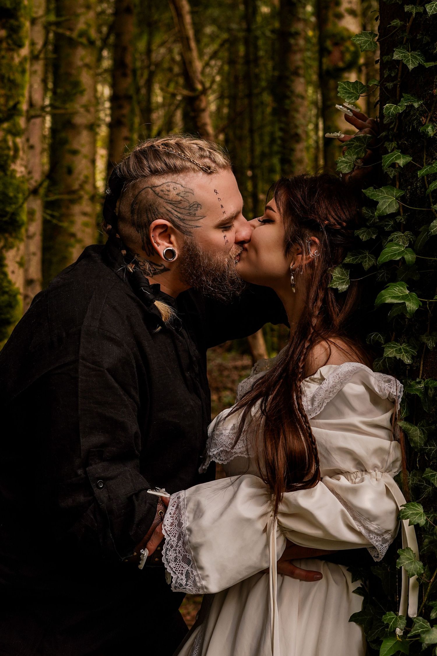 Tattooed groom and bride sharing romantic kiss in enchanted forest wedding setting.