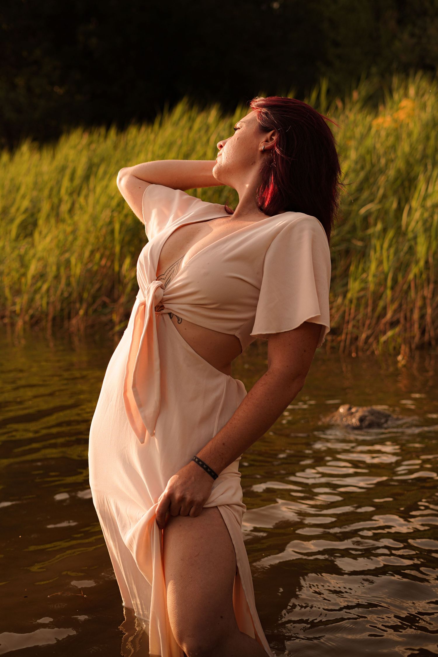 Woman in white tied dress stands in river at golden sunset.
