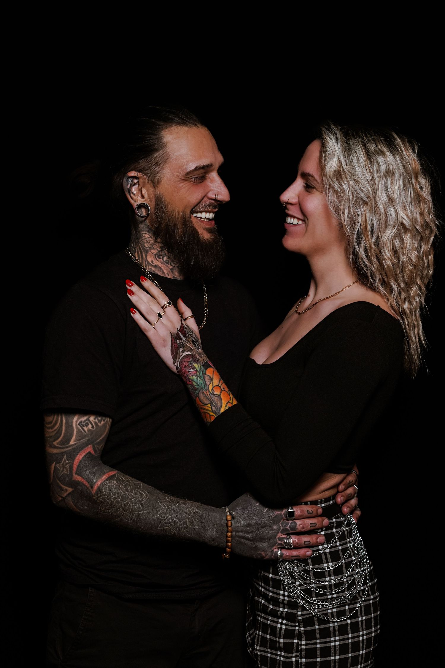 Smiling tattooed couple embracing and laughing together against a dark black background.