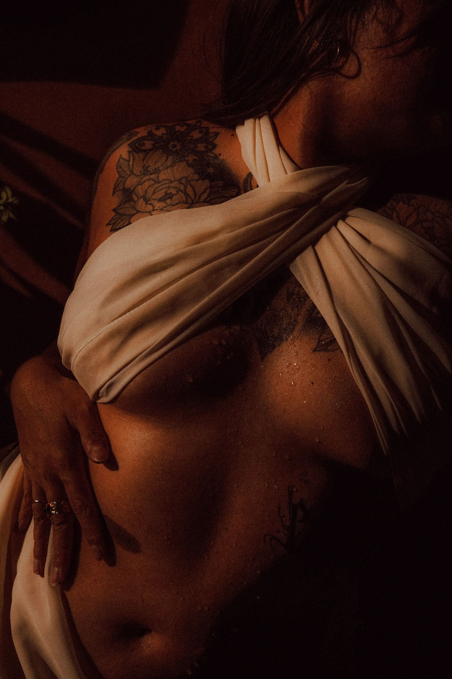Close-up boudoir shot of a tattooed torso wrapped in cream draped fabric in warm dark studio setting.