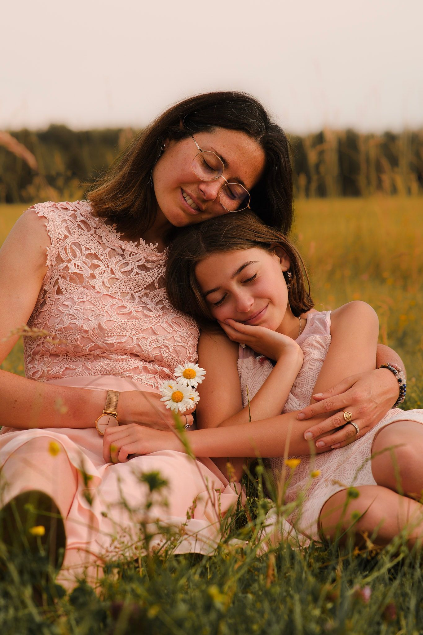 A mother and daughter sit together in a sunny meadow, sharing a warm embrace among wildflowers.