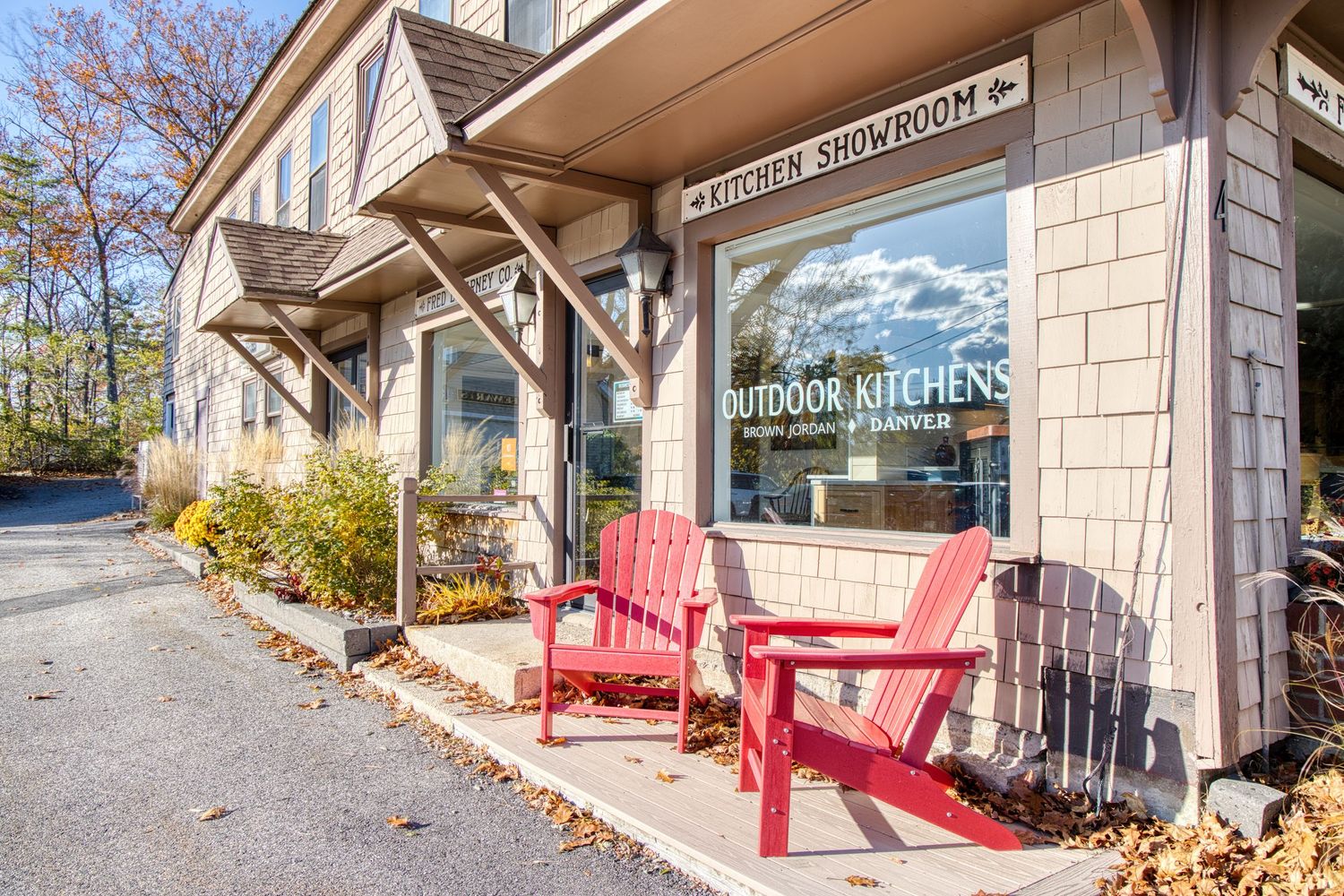 Red Adirondack chairs line the entrance of a stone building with autumn leaves scattered on the ground.