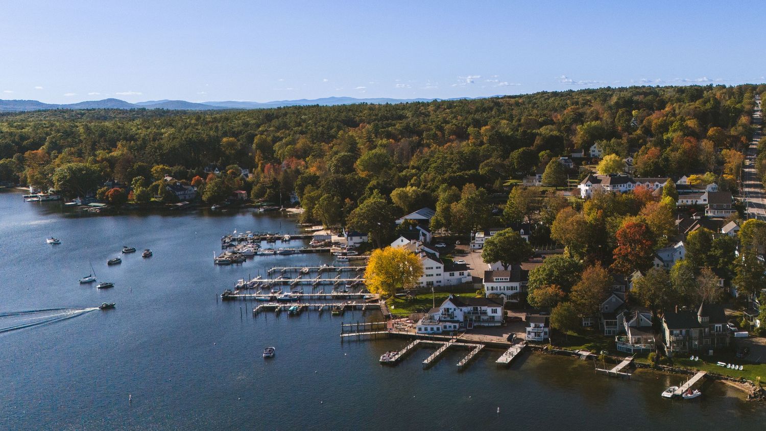 Aerial view of a coastal New England harbor with autumn foliage, boats, and docks along the shoreline.