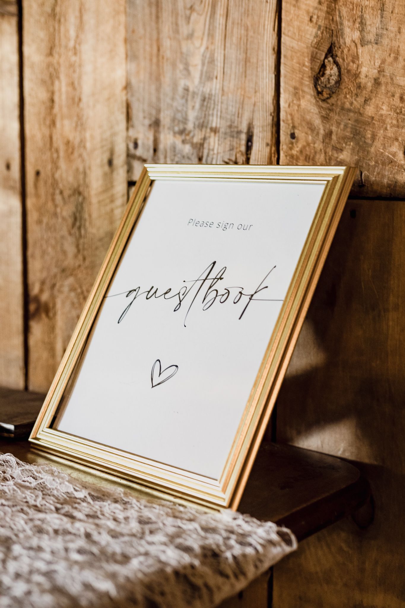 Gold framed guestbook sign with handwritten text sits on rustic wooden surface with textured fabric.