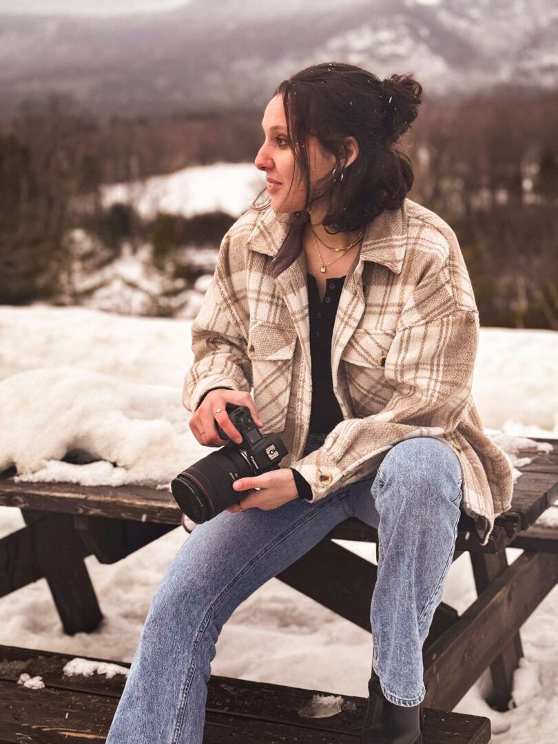 A photographer in a plaid shearling jacket and jeans sits on a snowy bench holding a camera with mountains in the background.