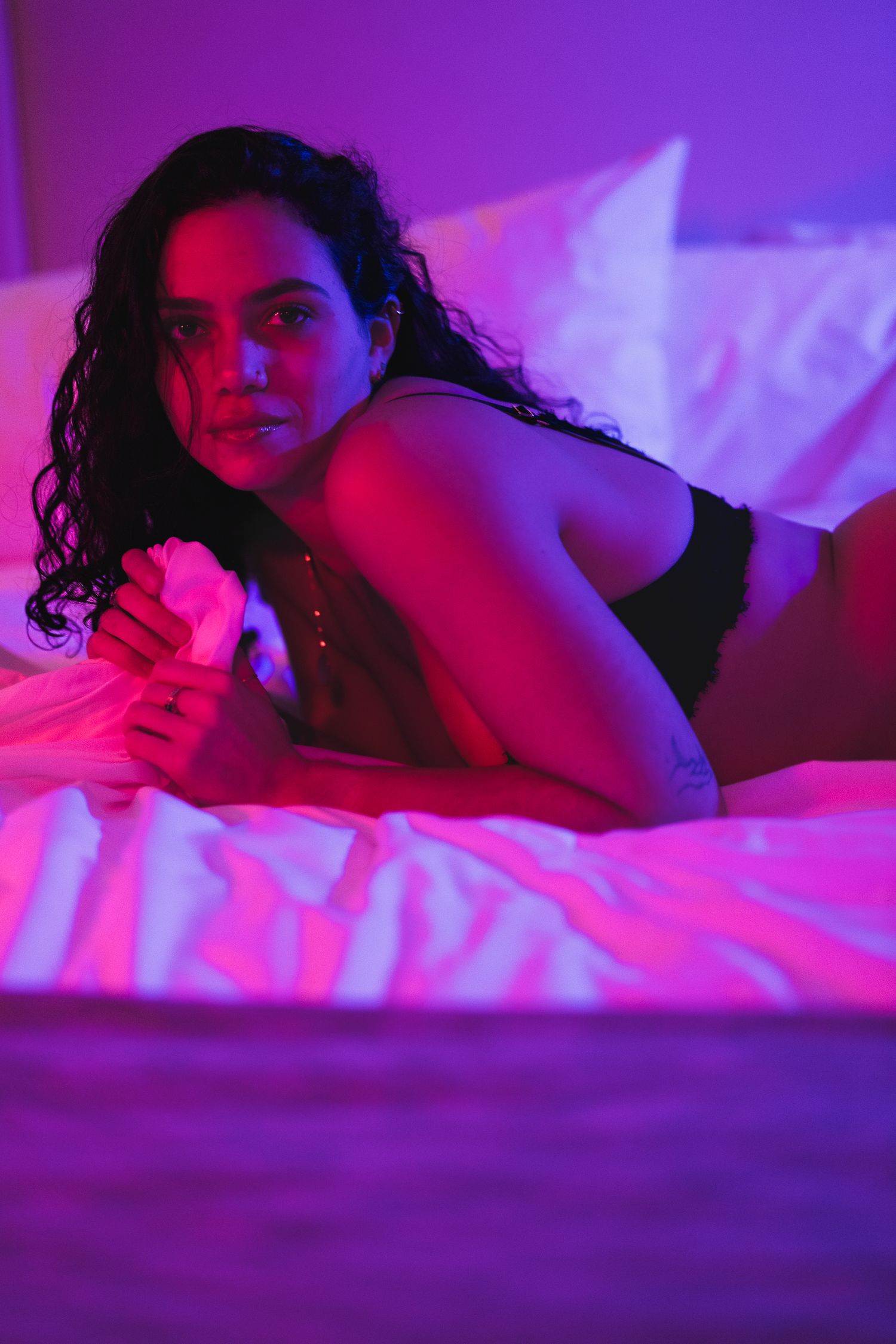 A boudoir photoshoot features dramatic purple and pink lighting on white bedsheets creating a sensual mood.