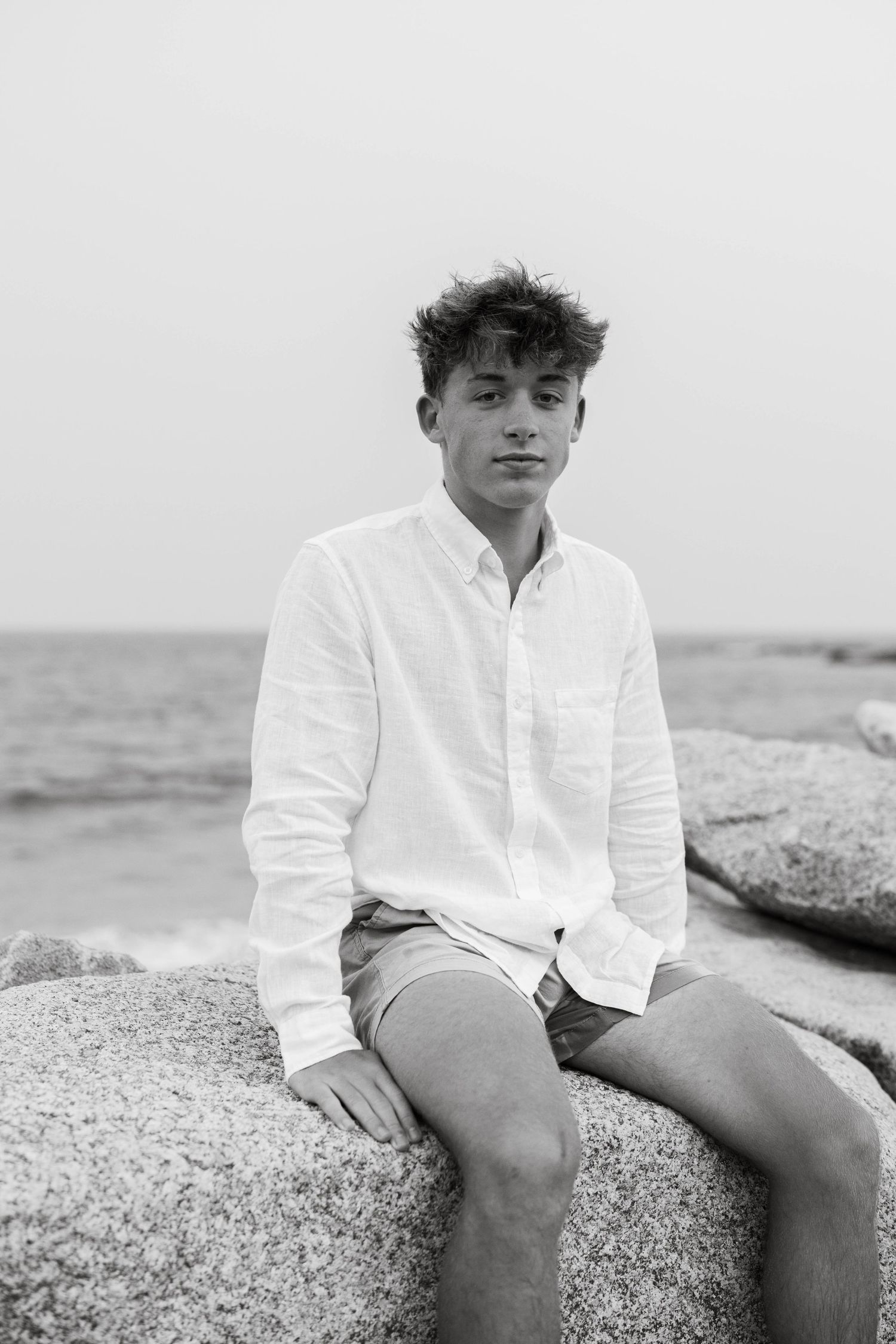 Black and white photo of person in white button-down shirt sitting on coastal rocks by the ocean.