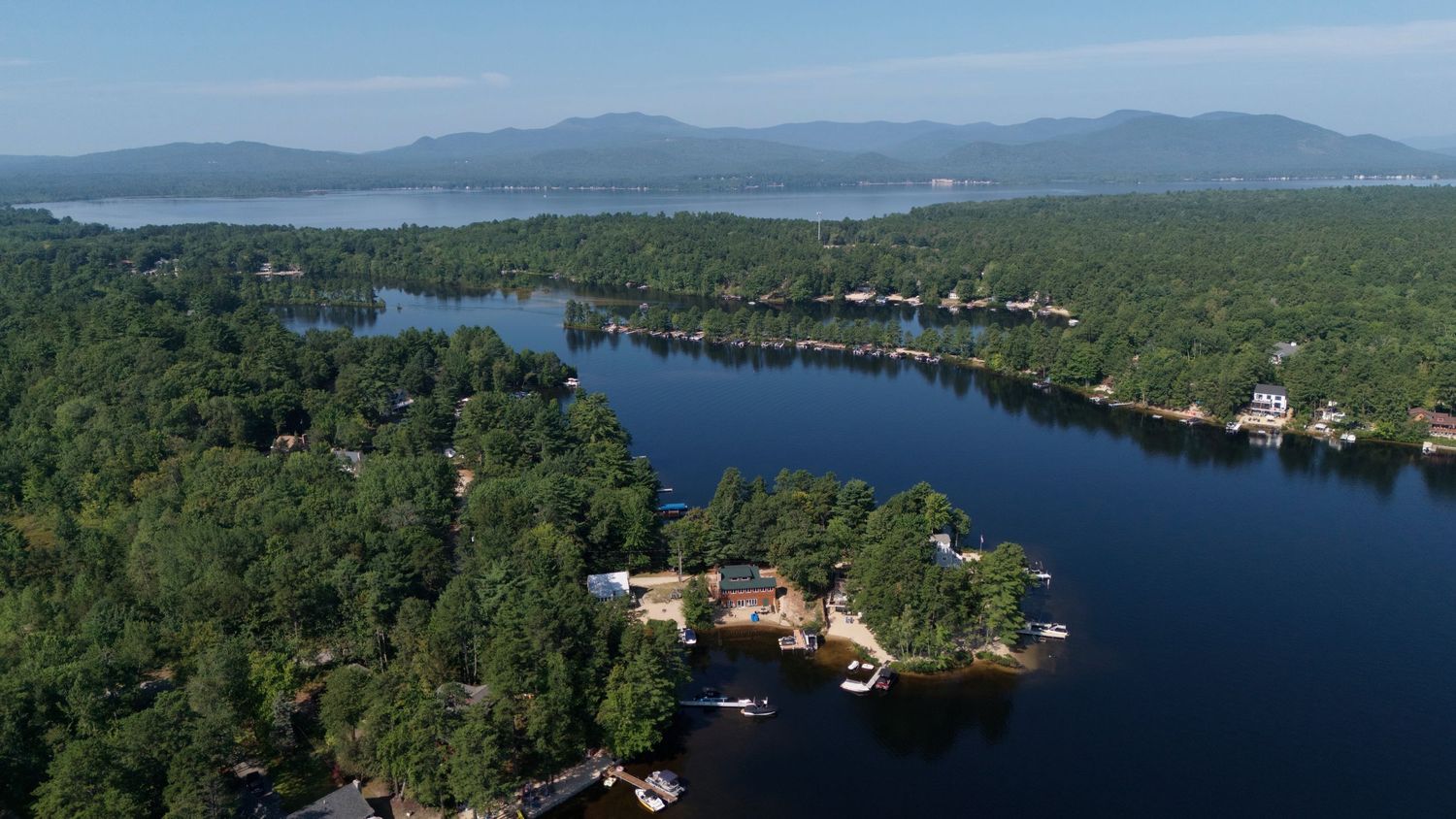 Aerial view of Lake Winnipesaukee shoreline with dense forest, waterfront homes, and distant mountains in New Hampshire.