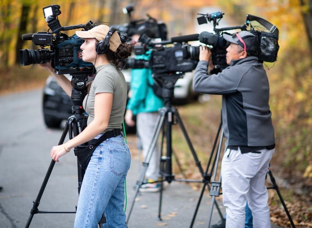 Professional camera operators with video equipment filming outdoors during autumn with colorful foliage in background.