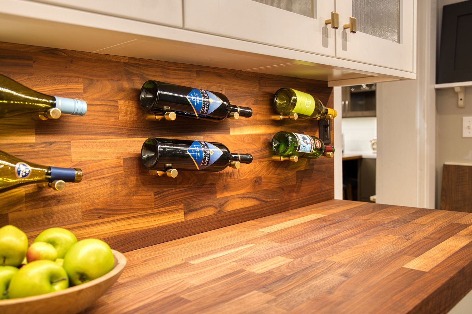 Modern kitchen counter with wood surface features mounted wine bottles and fresh green apples in a bowl.