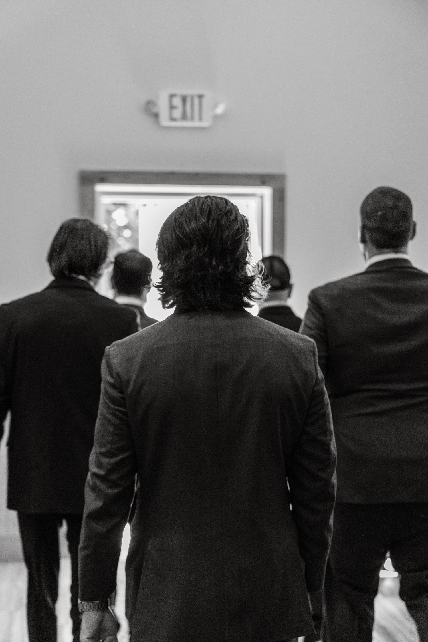 A black and white photo shows a line of people wearing dark suits viewed from behind while standing in a row.