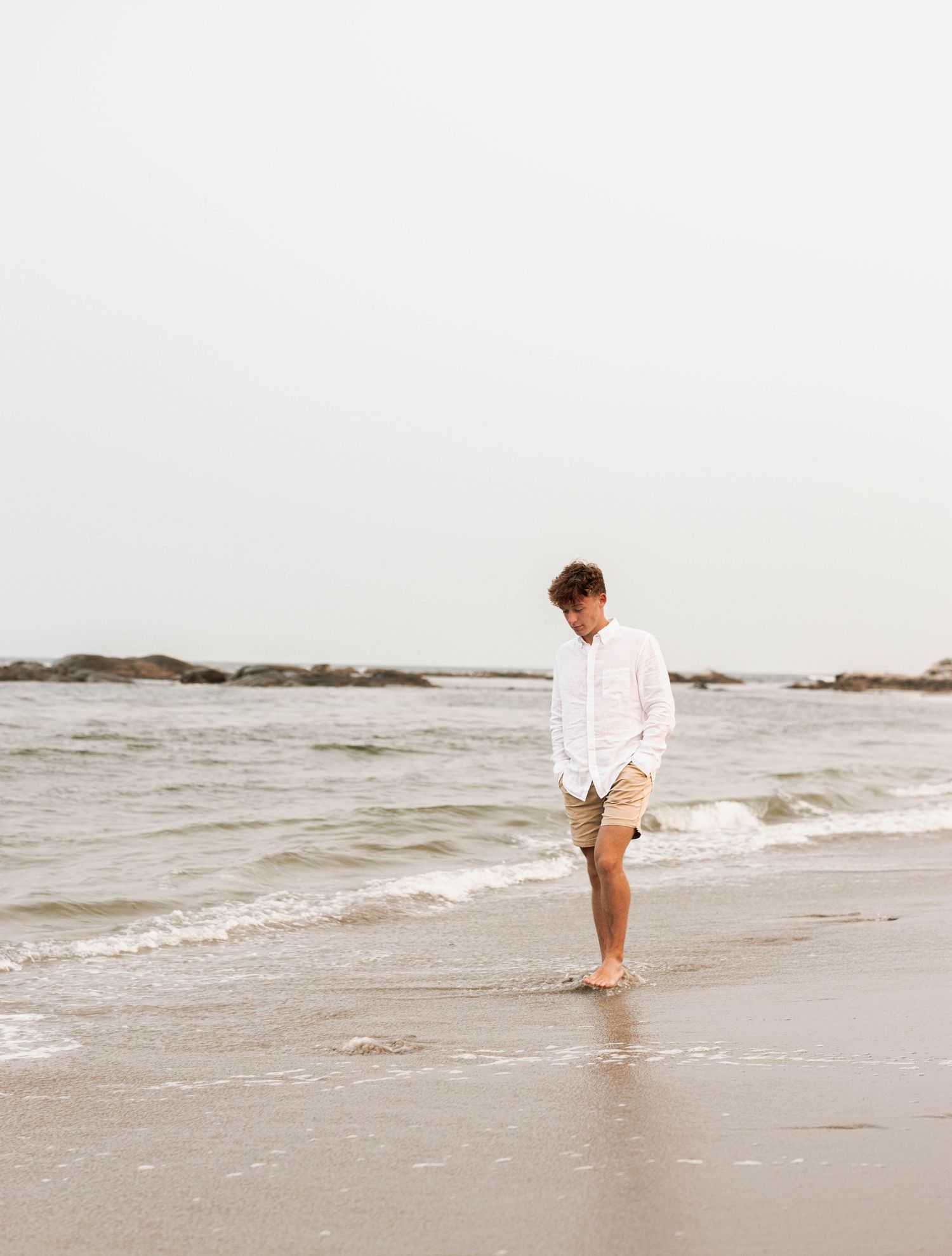 Person in white shirt and khaki shorts standing alone on misty beach shoreline looking down at waves.
