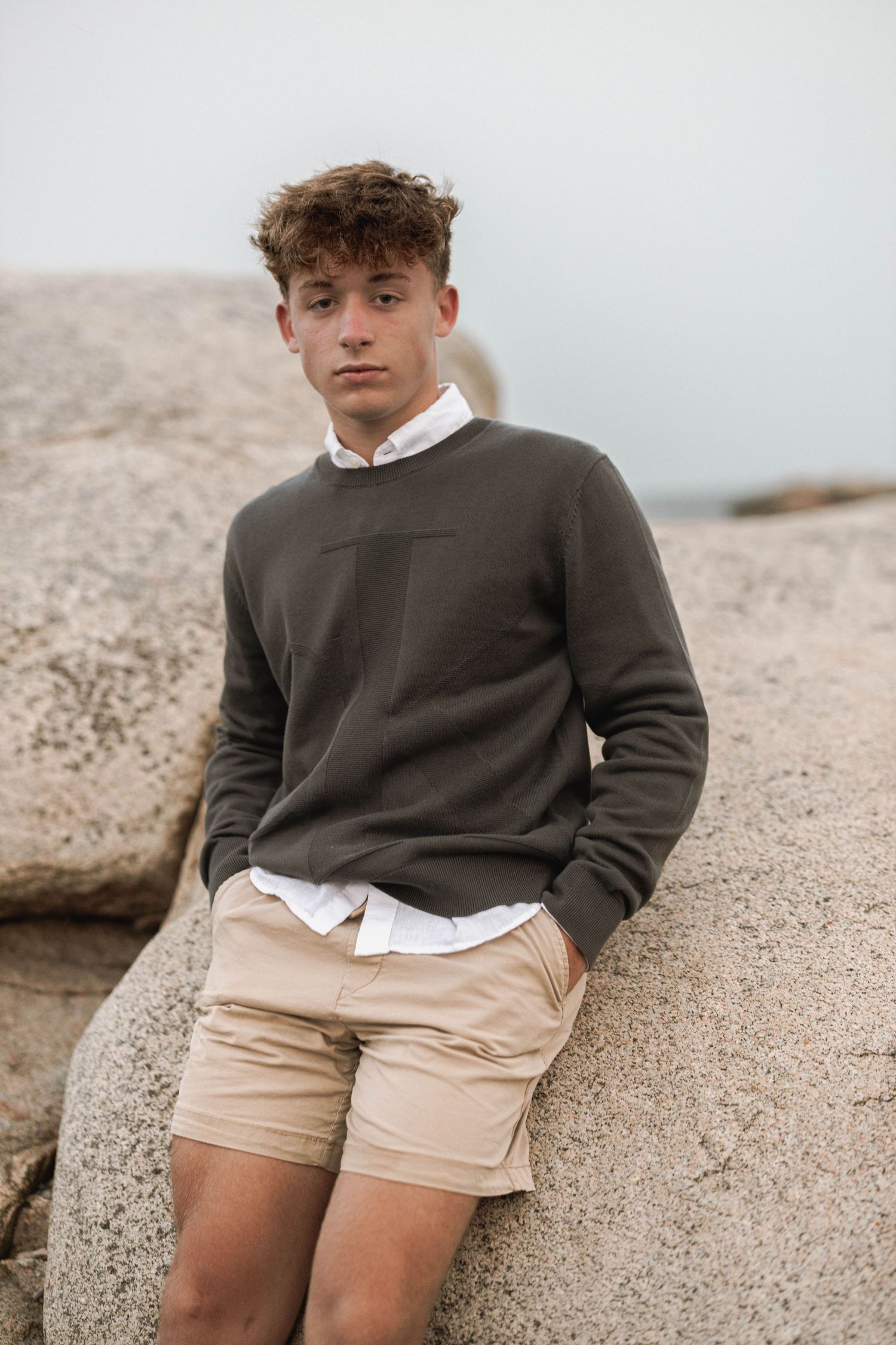 A person wearing a gray sweater over white shirt and khaki shorts poses against a rocky coastal backdrop.