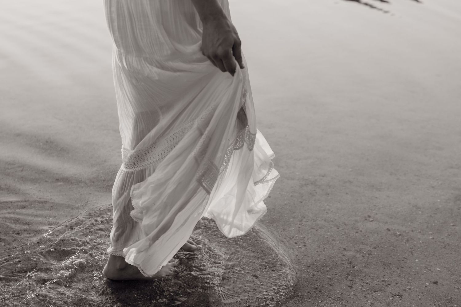 A couple in wedding attire walks along a misty beach shoreline with their reflections visible in the wet sand.