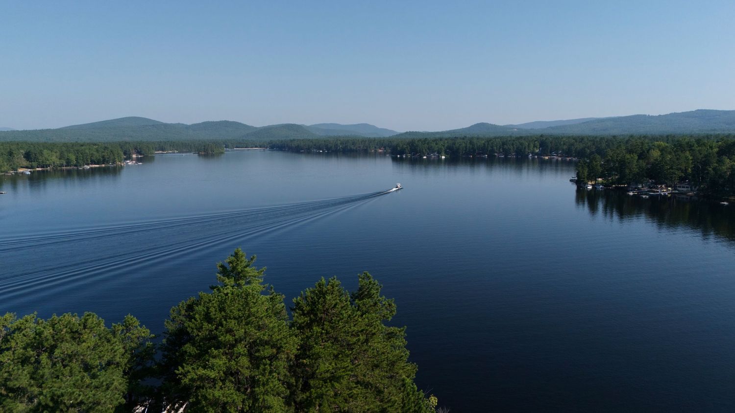 Panoramic view of a serene lake surrounded by green mountains and forests on a sunny summer day.