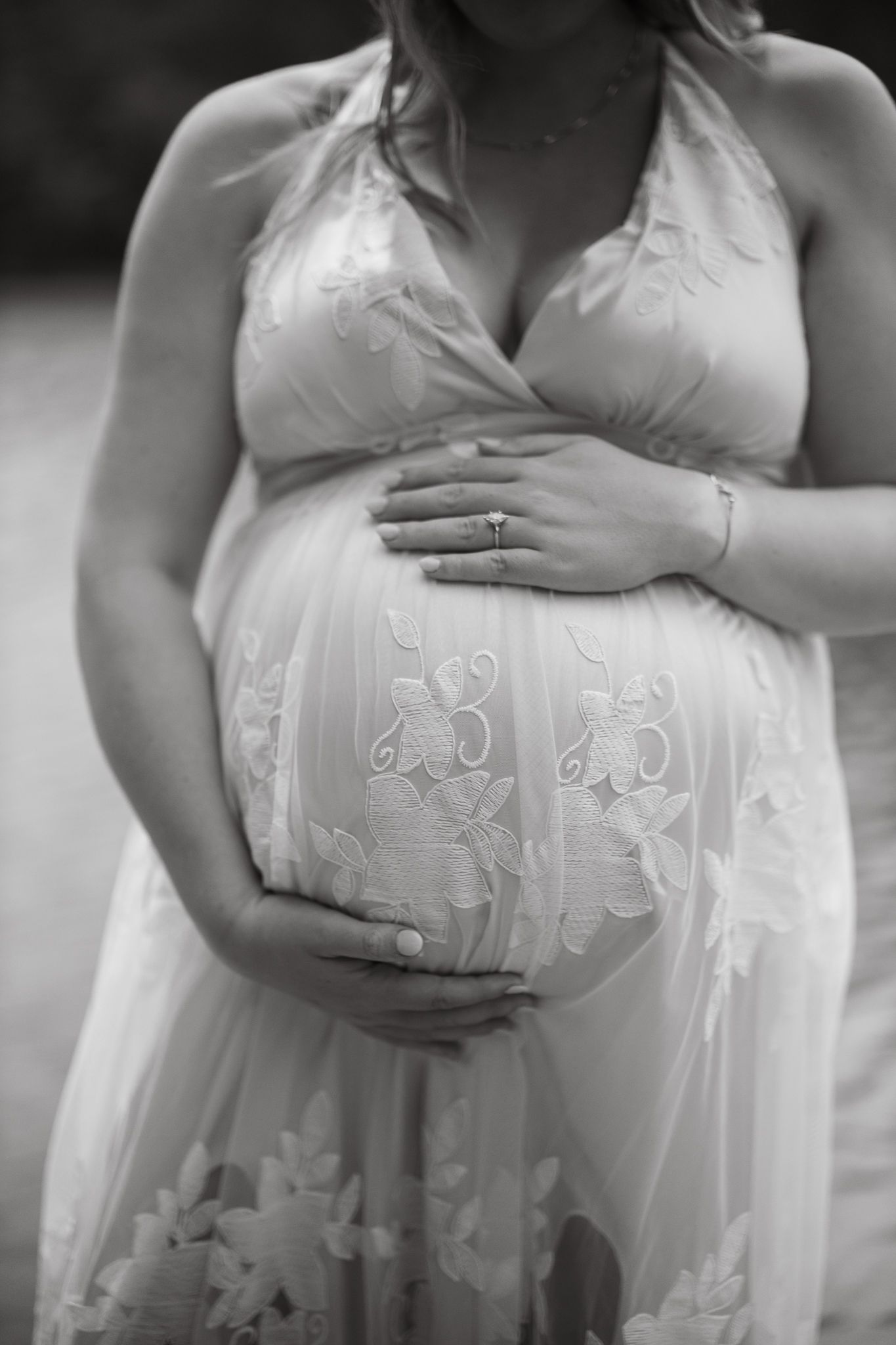Black and white maternity photo showing a pregnant belly in an embroidered white dress with hands cradling the bump.
