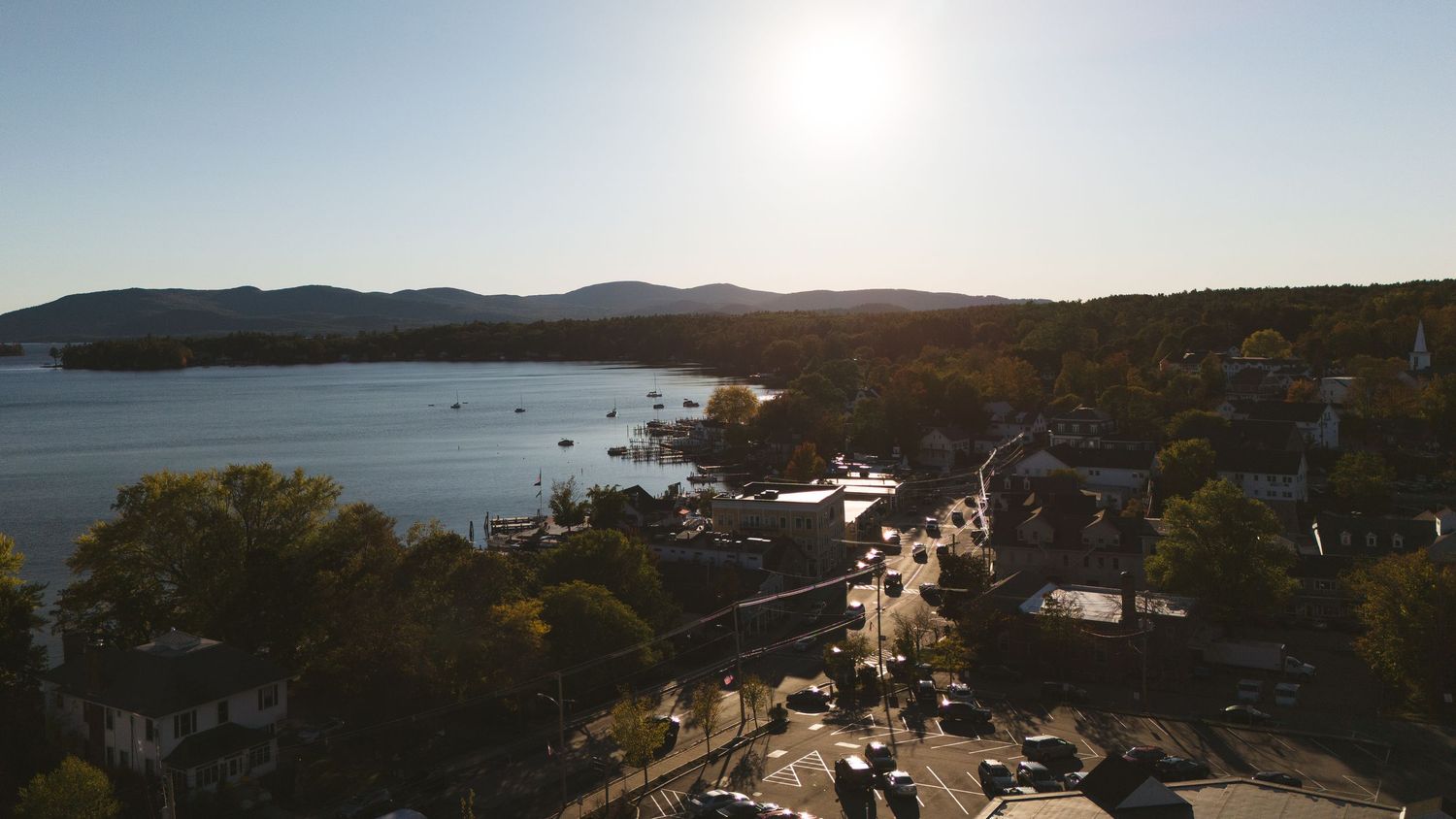 Panoramic sunset view of a coastal town with mountains, boats in the harbor, and autumn trees along the shoreline.