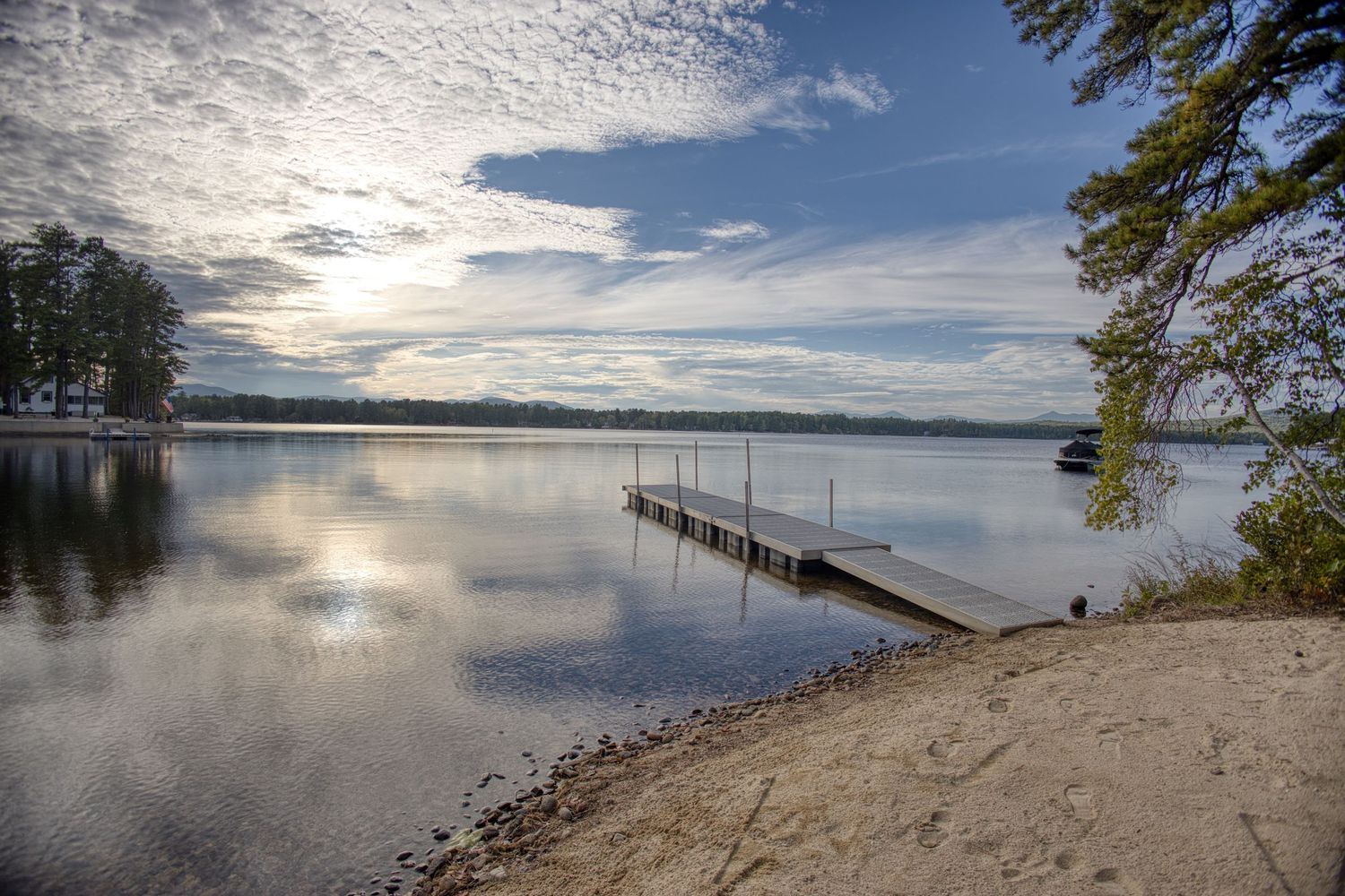Panoramic view of a calm lake with sandy shoreline and distant trees reflecting in the tranquil waters on a sunny day.