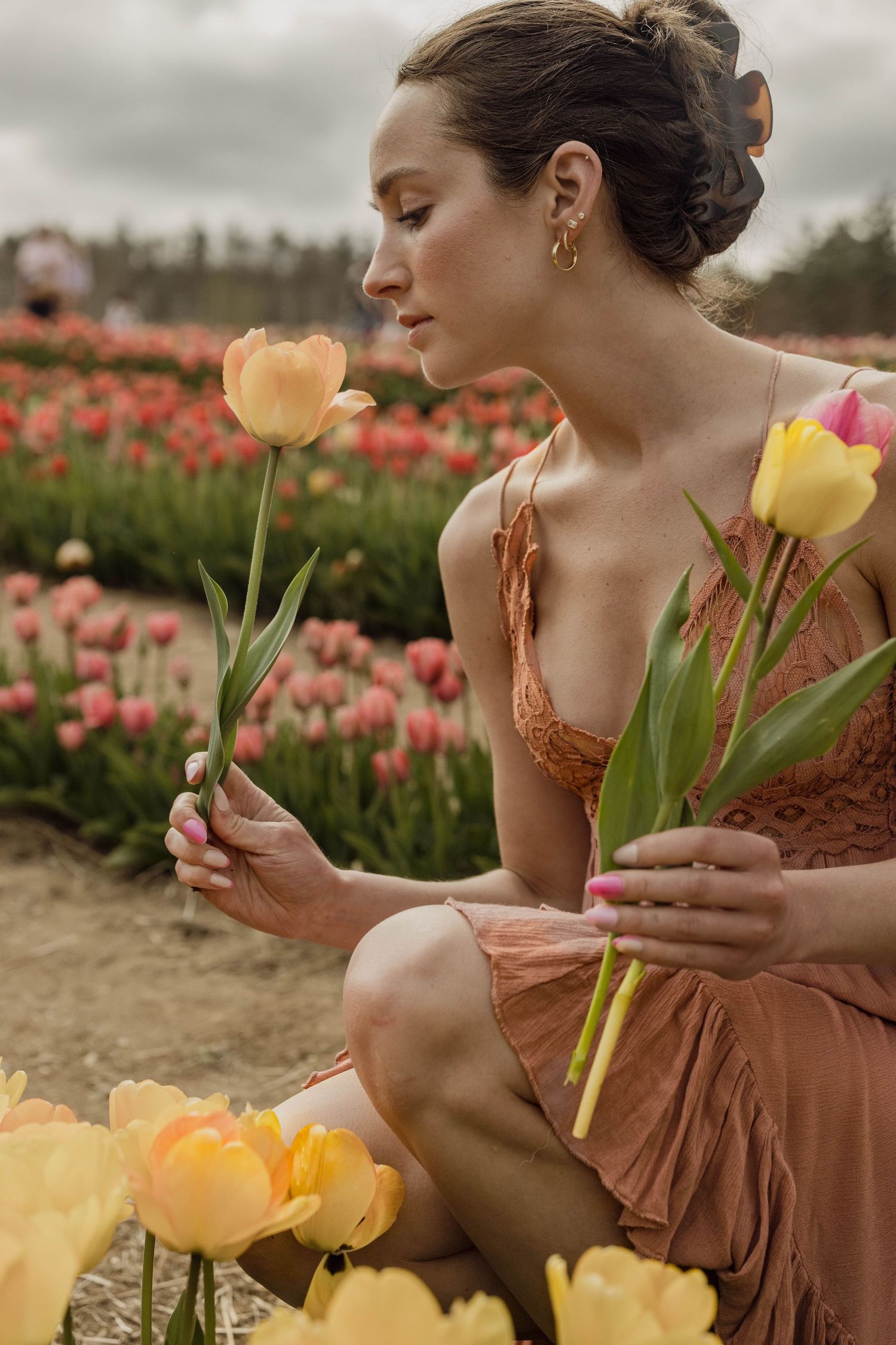 A person in a peach-colored dress admires tulips while sitting in a flower field on a cloudy day.