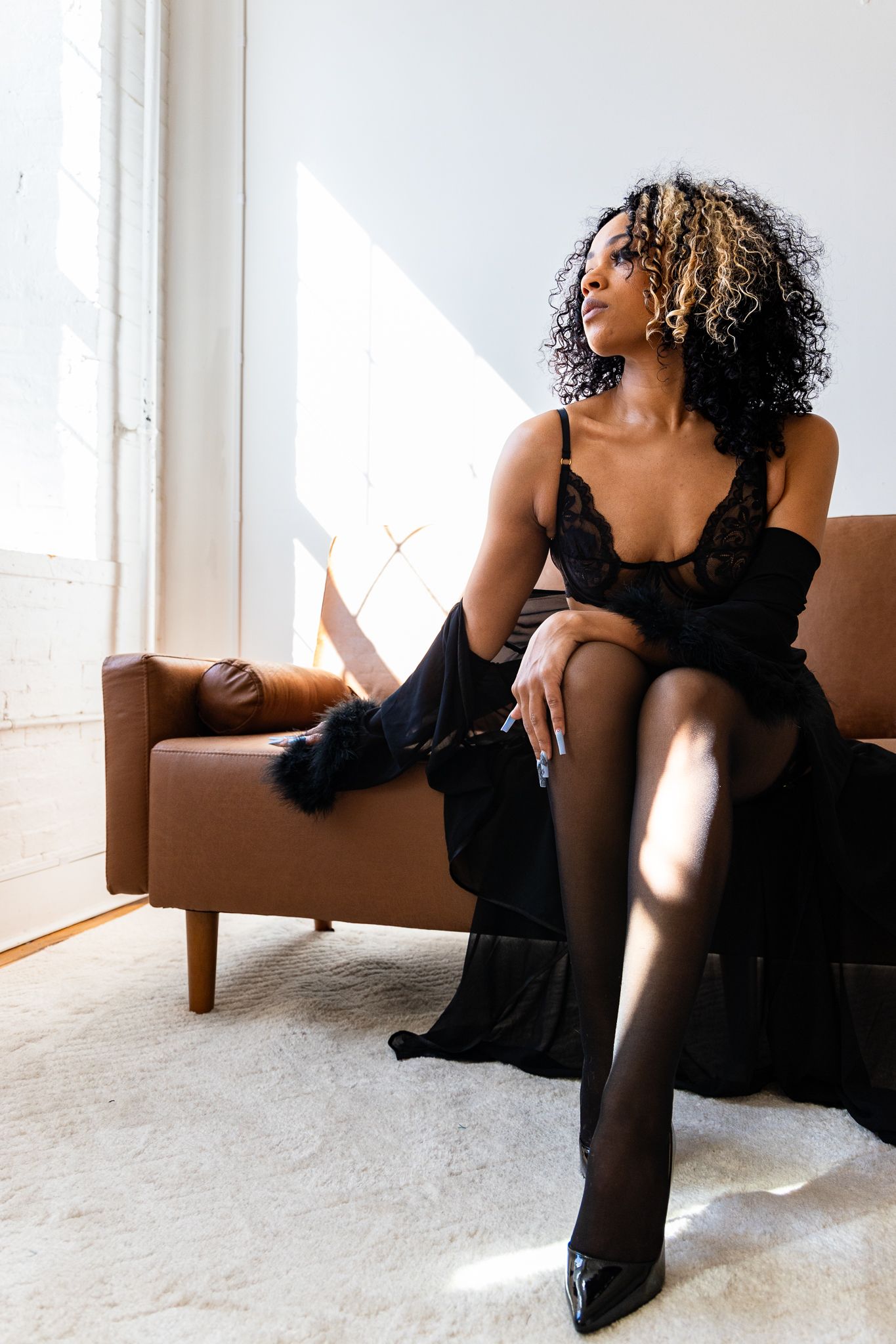 Woman in elegant black dress poses on brown leather chair in sunlit room with white walls and carpet.