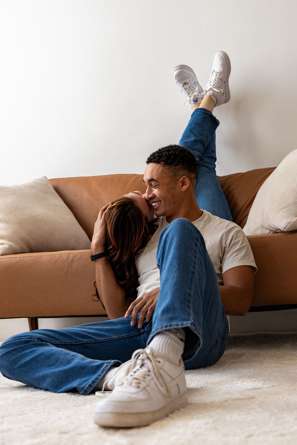 A person in blue jeans and white sneakers lies casually on a beige carpet next to a brown couch.