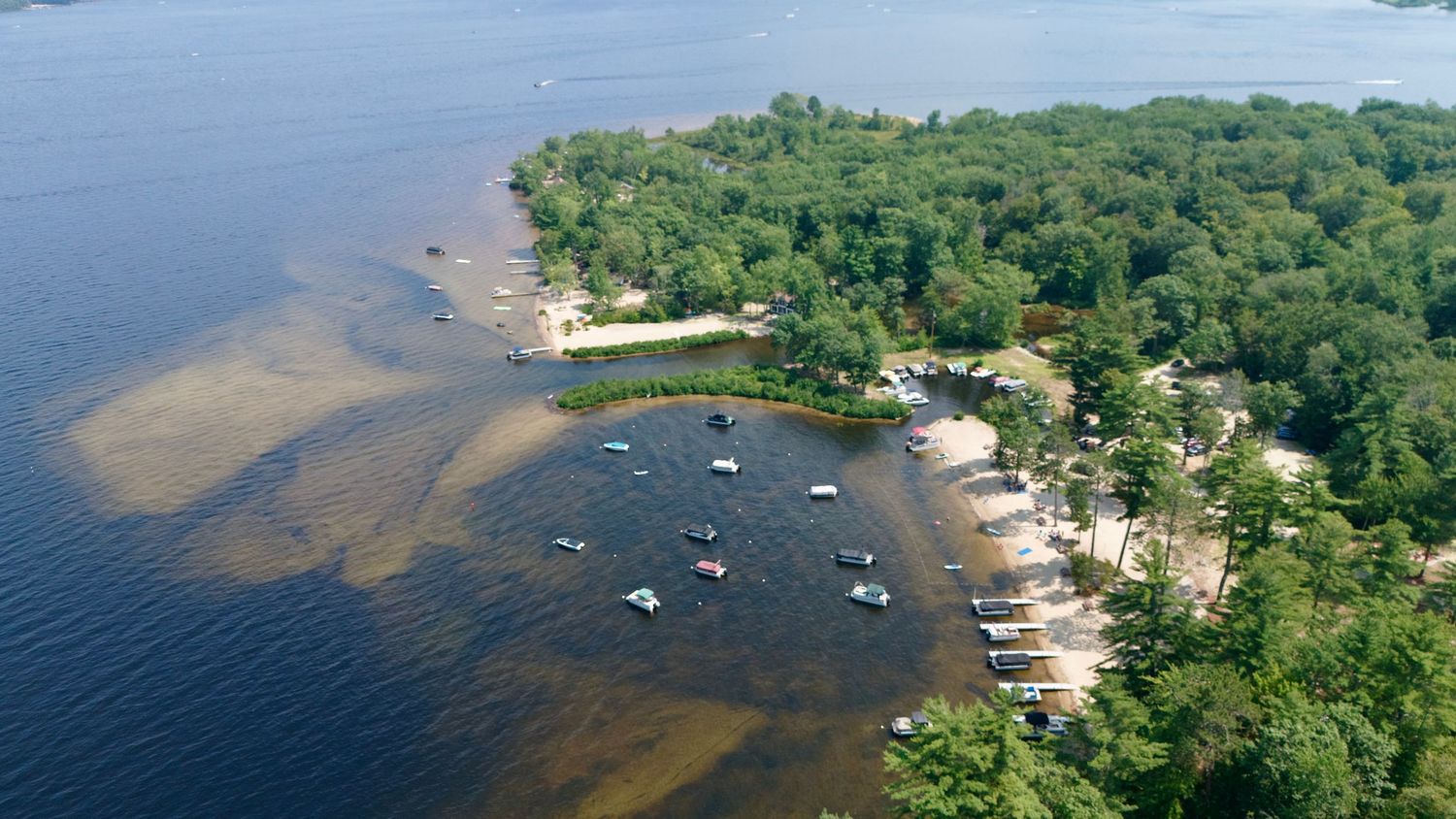 Aerial panoramic view of boats anchored along a tree-lined lakeshore with clear blue waters extending into the distance.