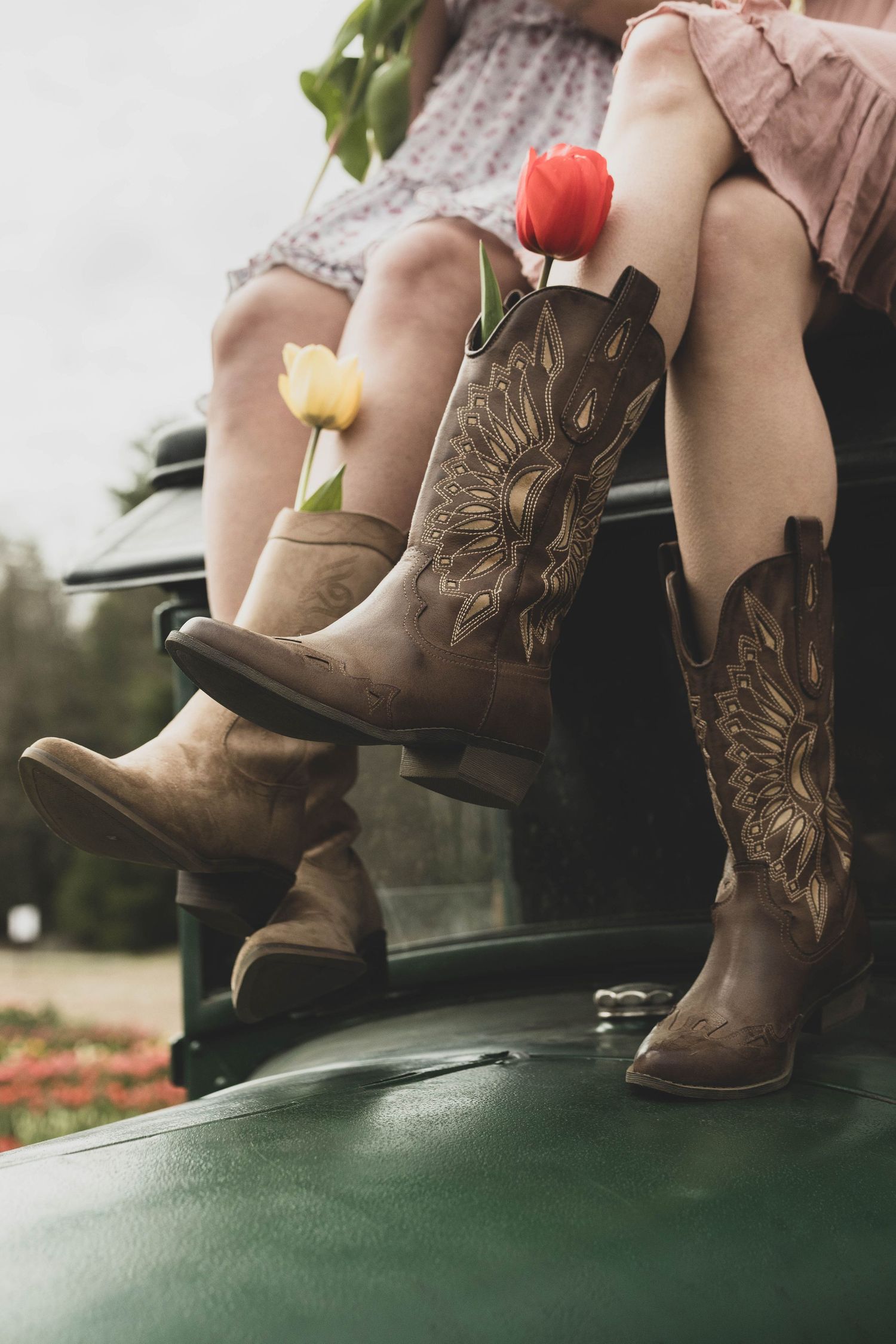 Brown leather cowboy boots with embroidered details rest beside delicate flowers on a green textured surface.