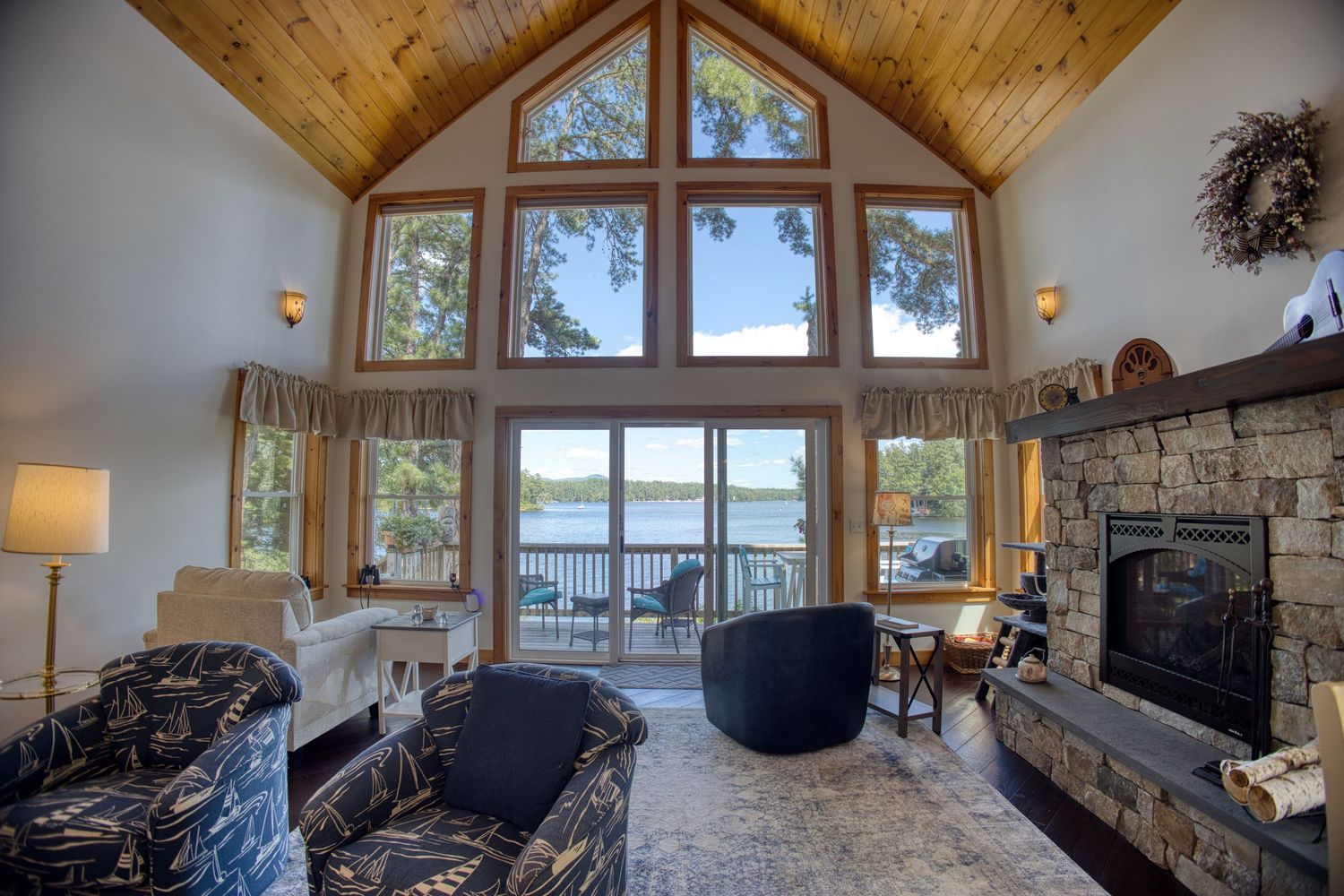 Luxurious lake house living room with vaulted wood ceiling, stone fireplace and panoramic water views through large windows.