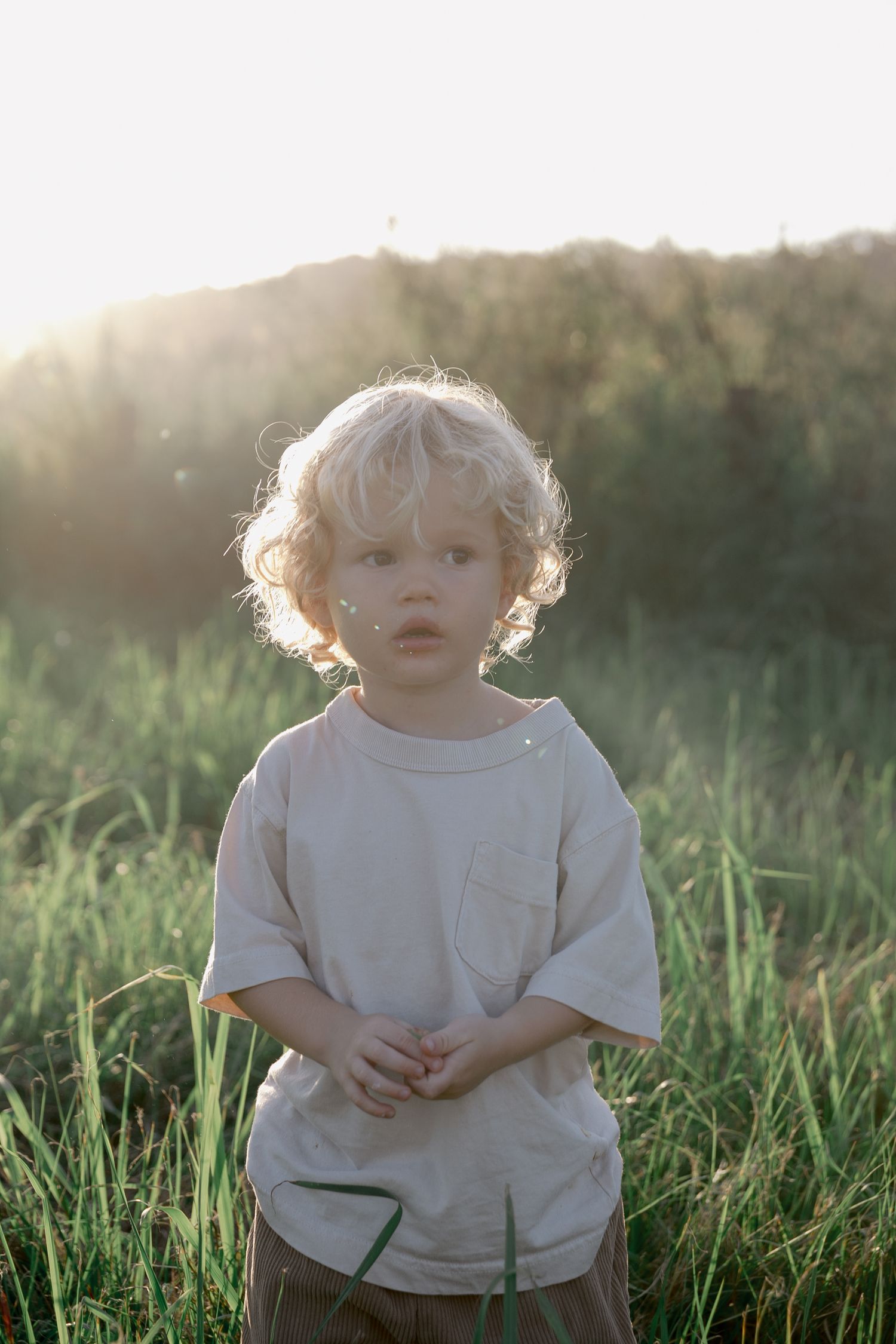 Curly blonde toddler in beige shirt stands in sunlit green field.
