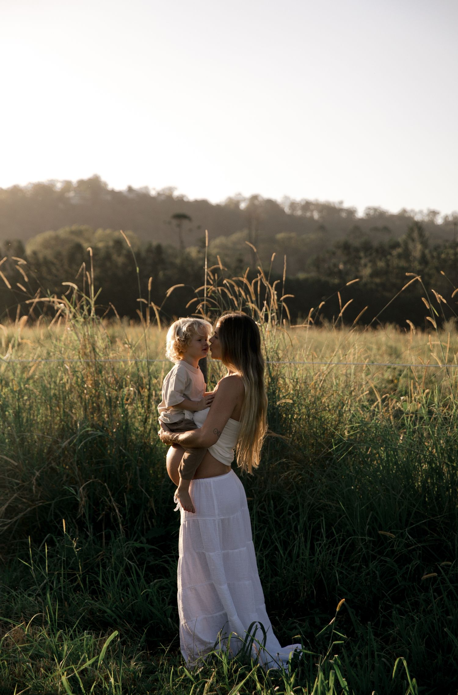 Mother in white dress holding and kissing toddler in golden sunlit grass field.