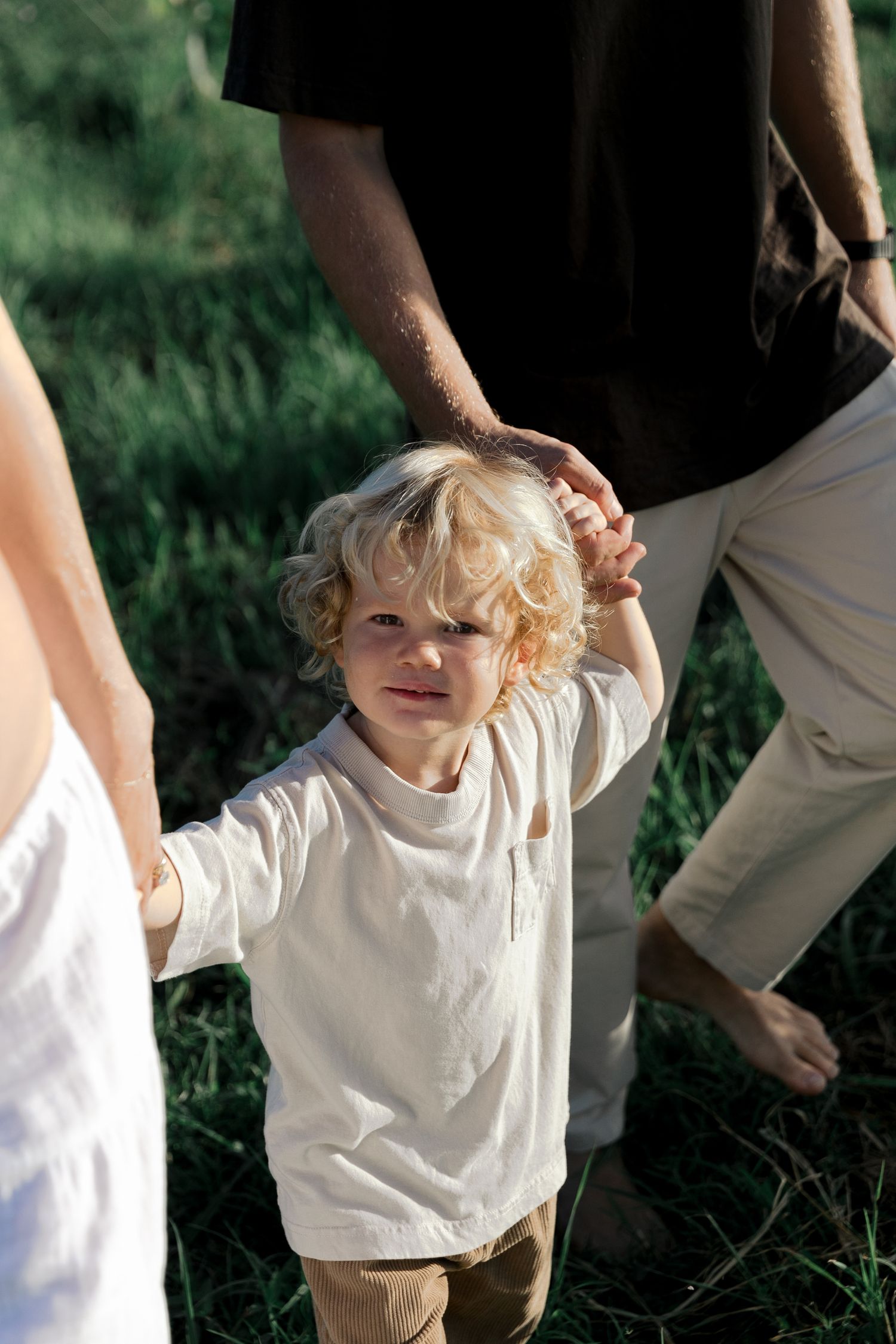Curly haired blonde toddler holding parents hands while walking in green meadow.