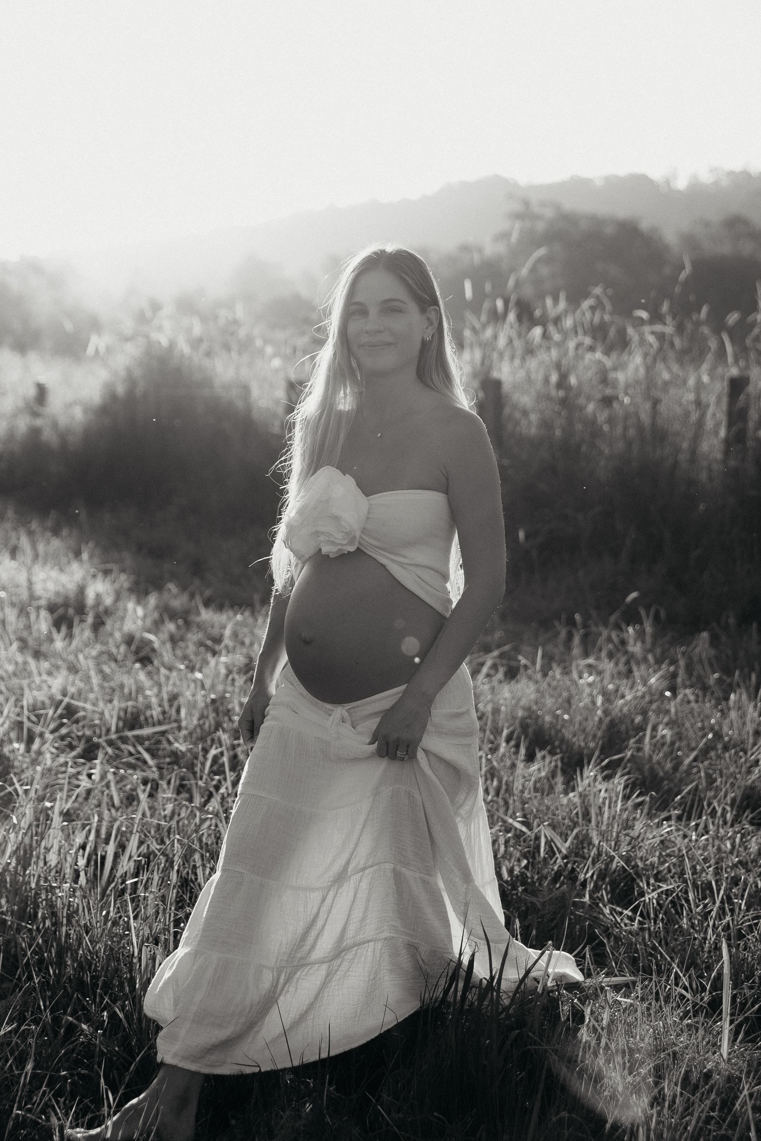 Black and white portrait of smiling pregnant woman in white dress in sunlit field.