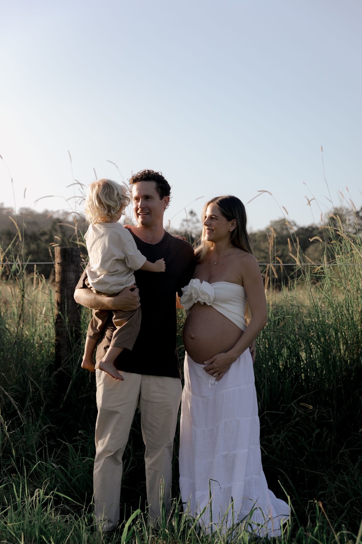 Smiling pregnant woman in white dress with husband holding toddler in green field.