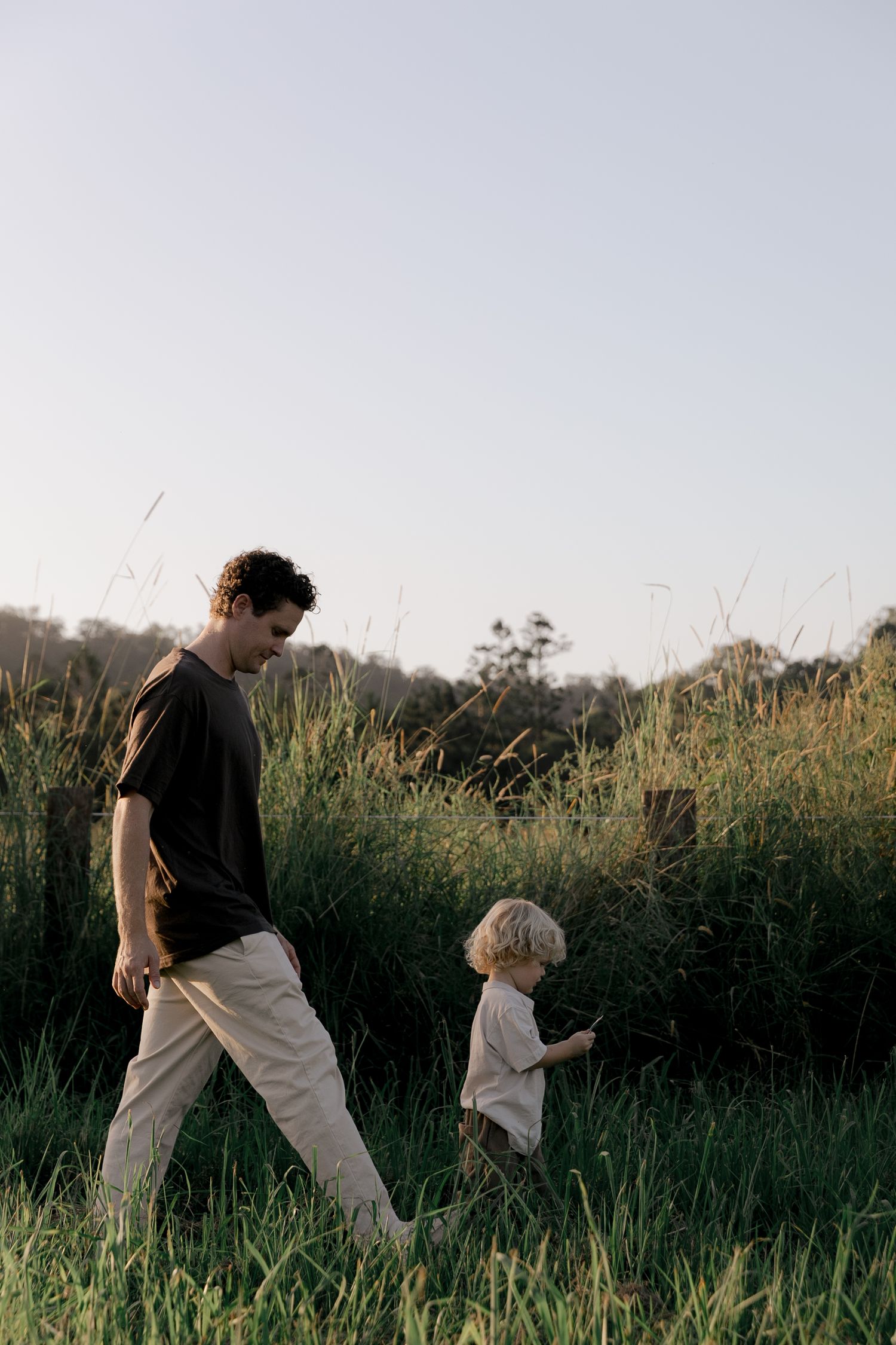 Father in black shirt walks with blonde toddler through tall green grass.