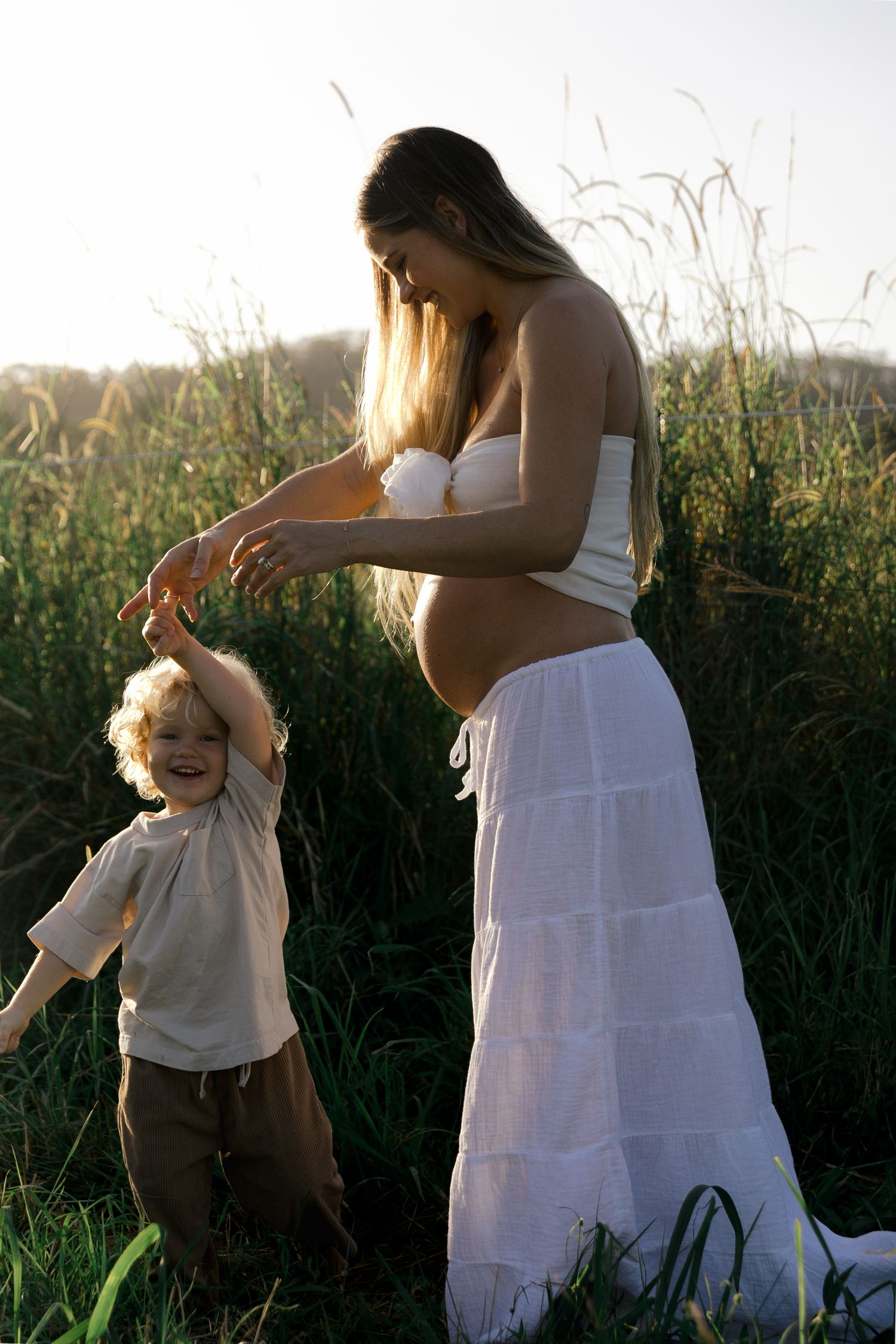 Pregnant woman in white skirt plays with curly-haired toddler in sunlit green field.