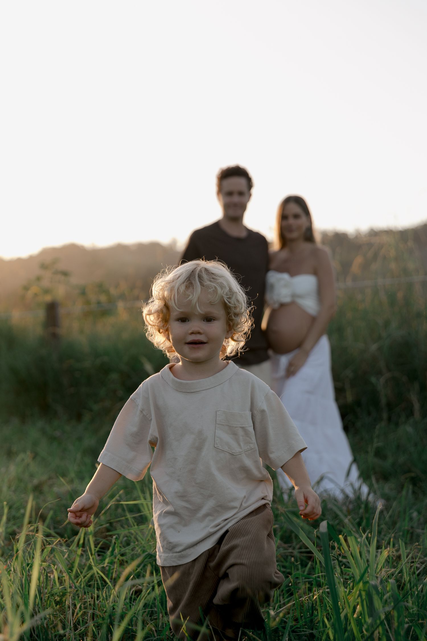 Curly-haired toddler runs toward camera with expecting parents in golden field.