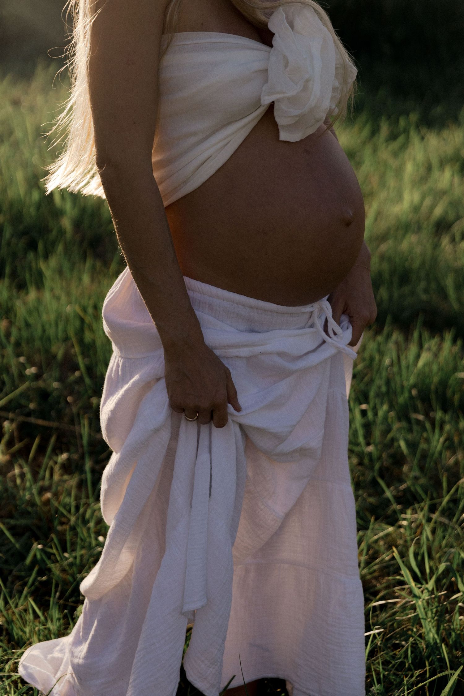 Close-up of pregnant woman's bare belly in white wrap skirt in field.