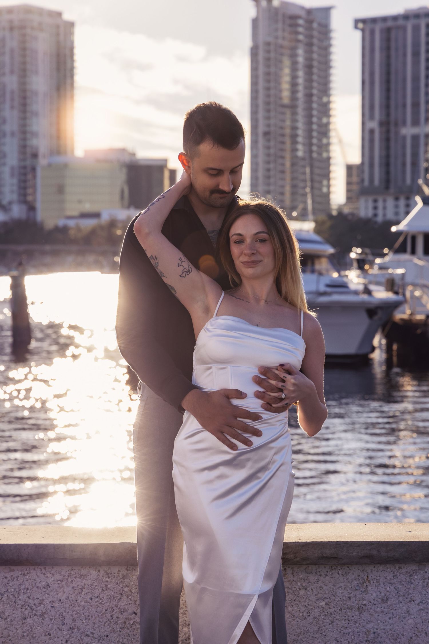 A romantic couple embraces at sunset by a marina with city skyscrapers in the background.