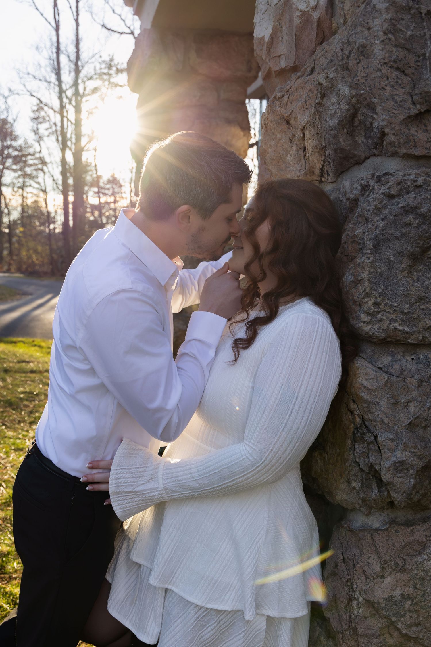 A romantic couple in white outfits sharing intimate moments against a tree trunk with sunlight filtering through branches.