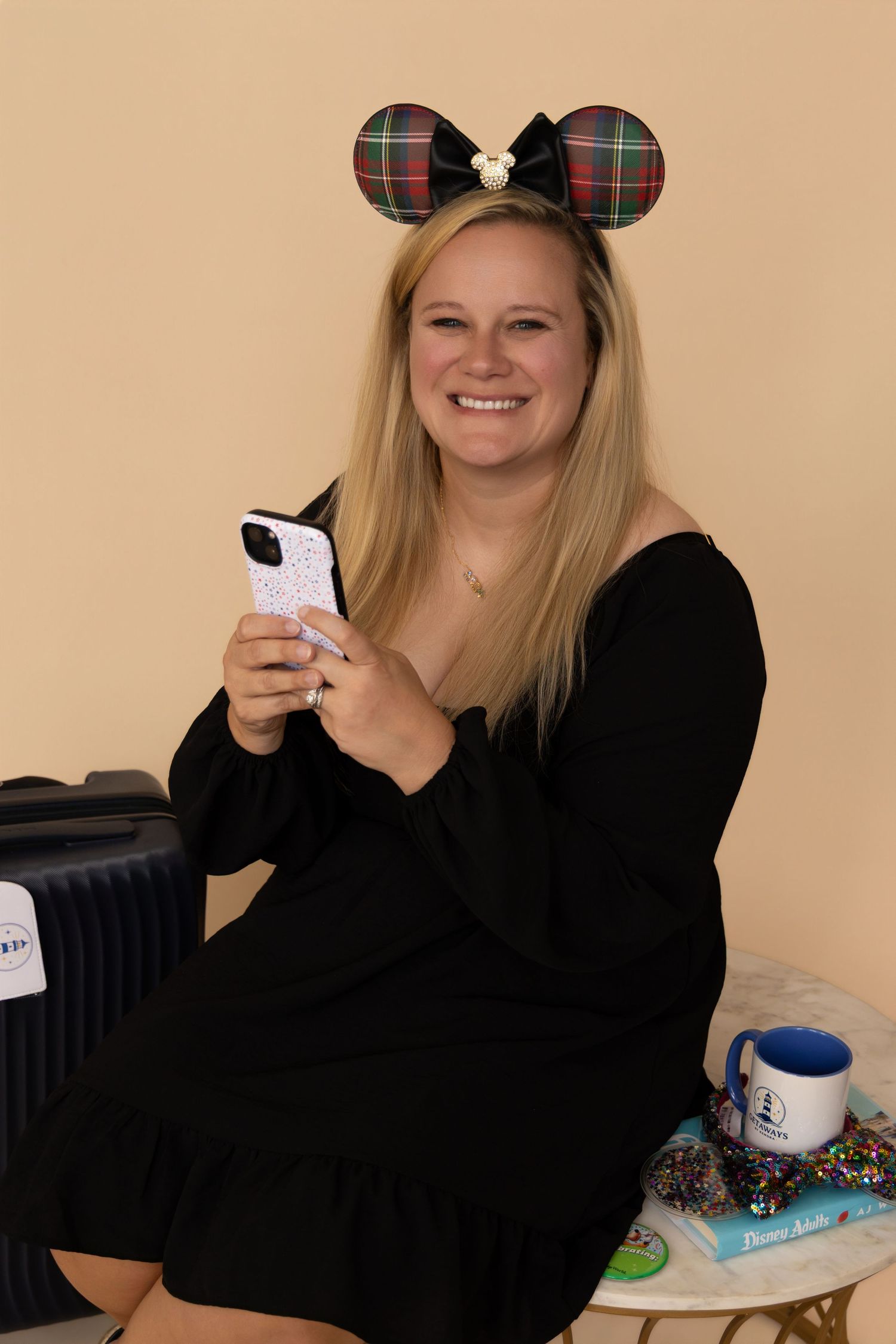 Smiling blonde woman wearing Minnie Mouse ears headband, black dress, holding phone, sitting near luggage and gifts.